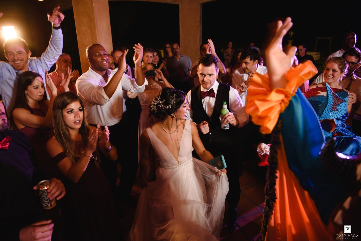 Bride and groom celebrate on the dance floor surrounded by cheering guests at a lively wedding reception.