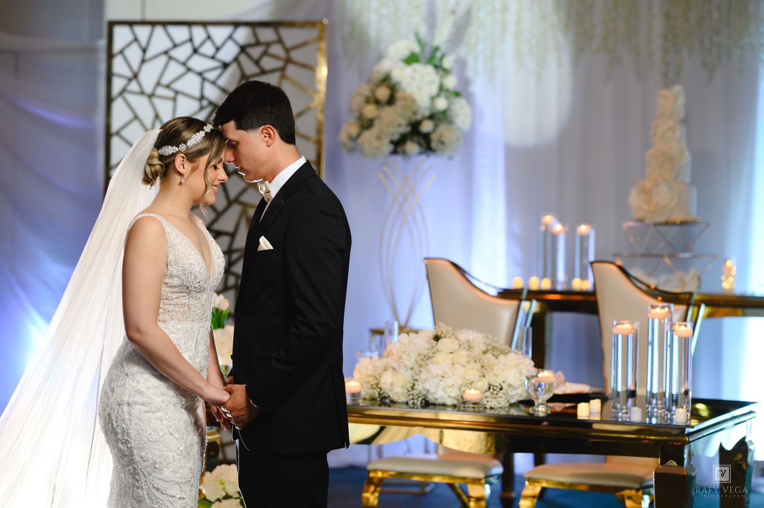 Bride and groom hold hands at elegant wedding altar with white floral arrangements, candles, and tiered cake backdrop.
