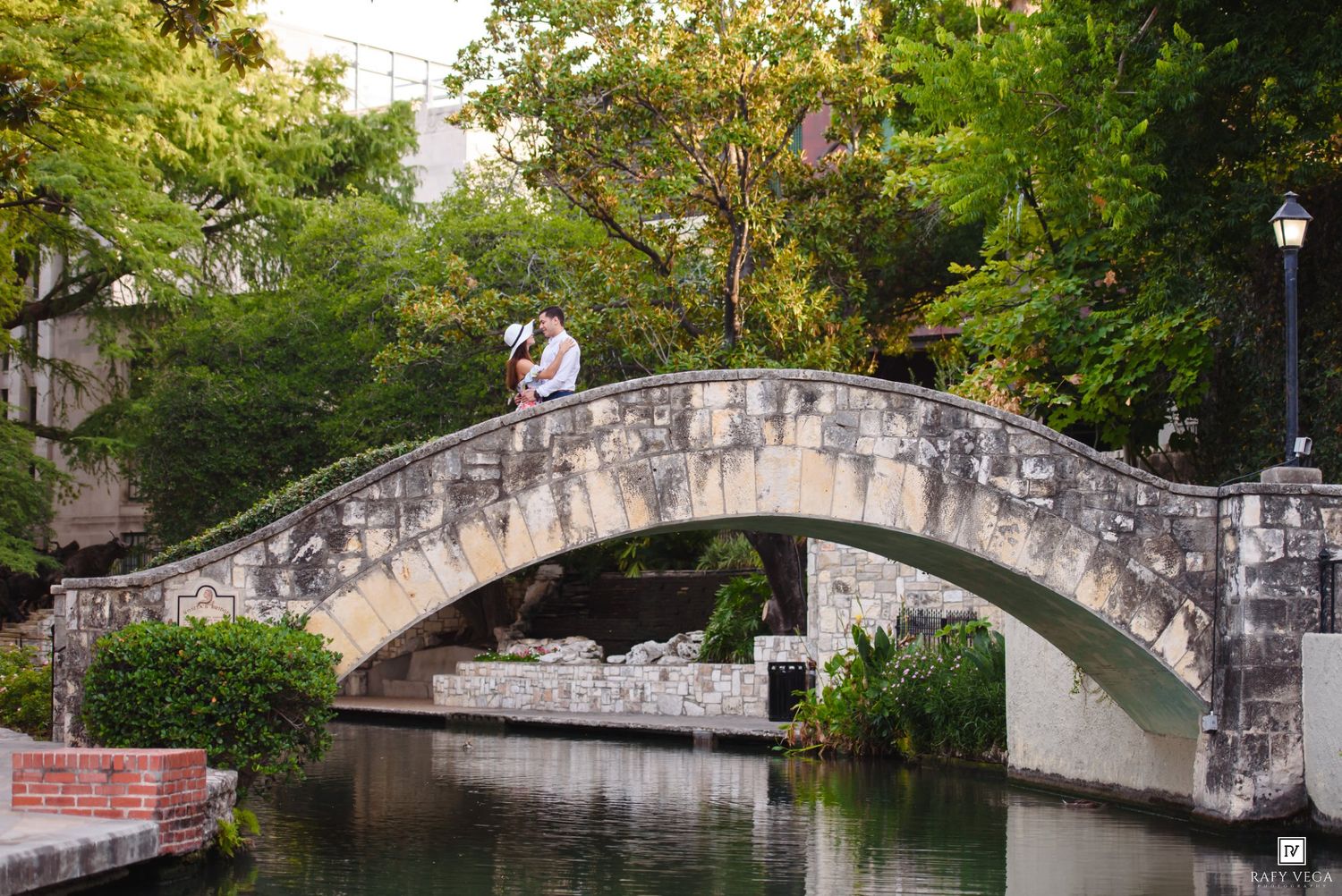 Couple embracing on a stone arch bridge over a calm river surrounded by lush green trees.