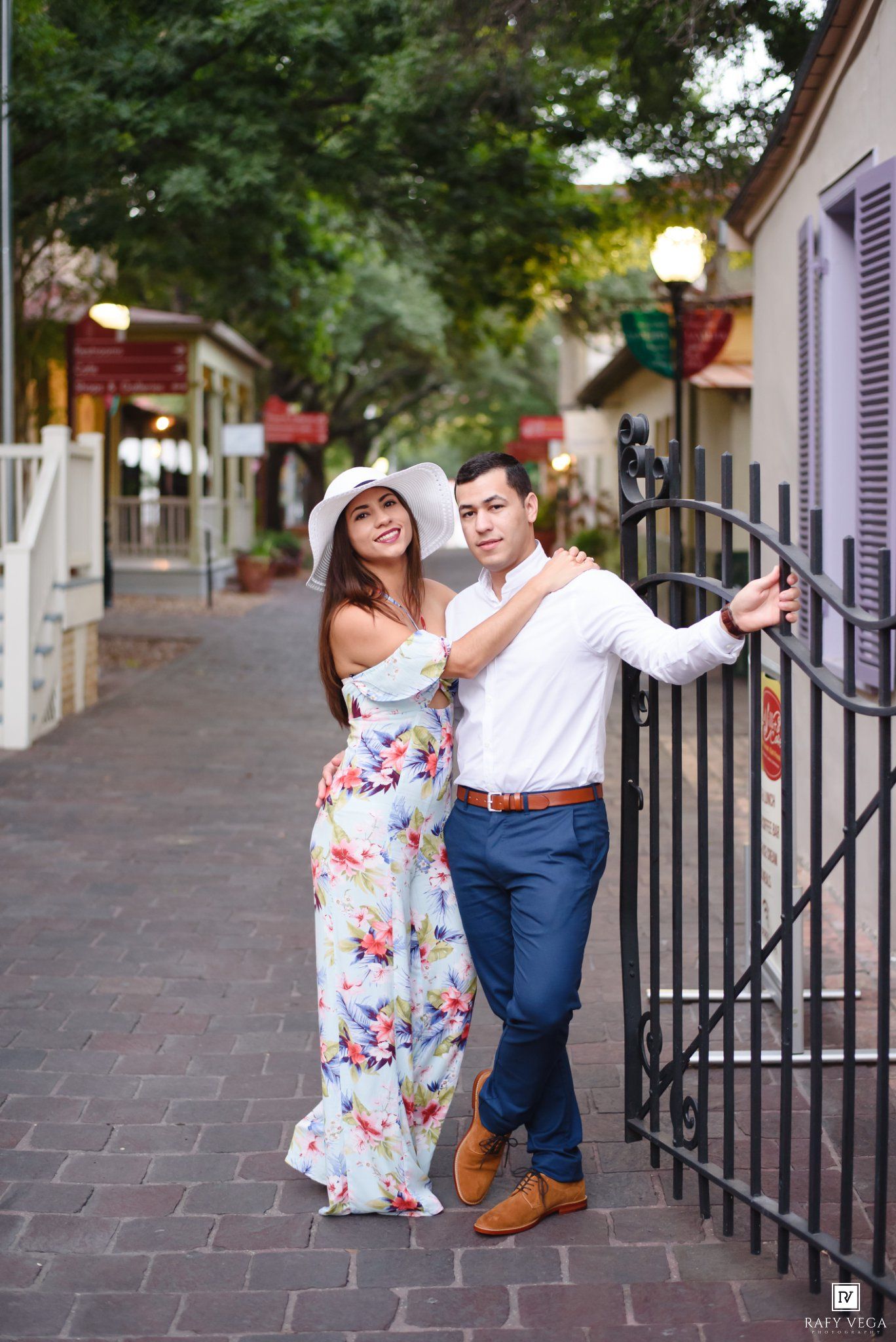 Couple posing by iron gate on brick walkway, woman in floral maxi dress and white hat, man in white shirt and navy pants.