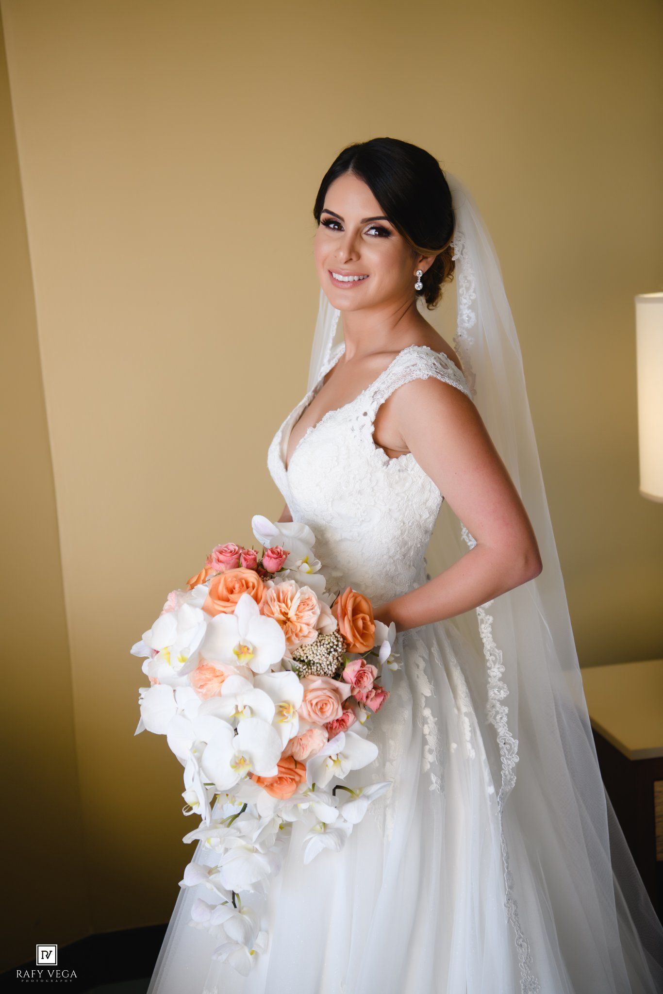 Bride in white lace gown holding cascading bouquet of white orchids and peach roses, wearing veil, smiling elegantly.