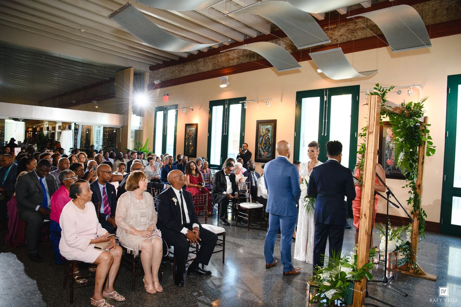 Elegant wedding ceremony in a decorated hall with guests seated, couple at altar adorned with greenery.