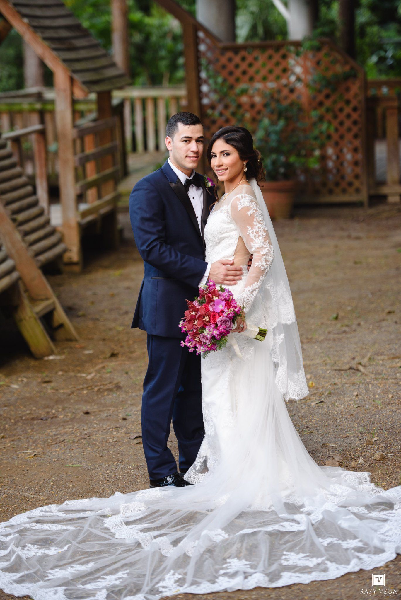 Bride in lace gown with long train holds pink bouquet beside groom in navy suit at outdoor wedding venue.