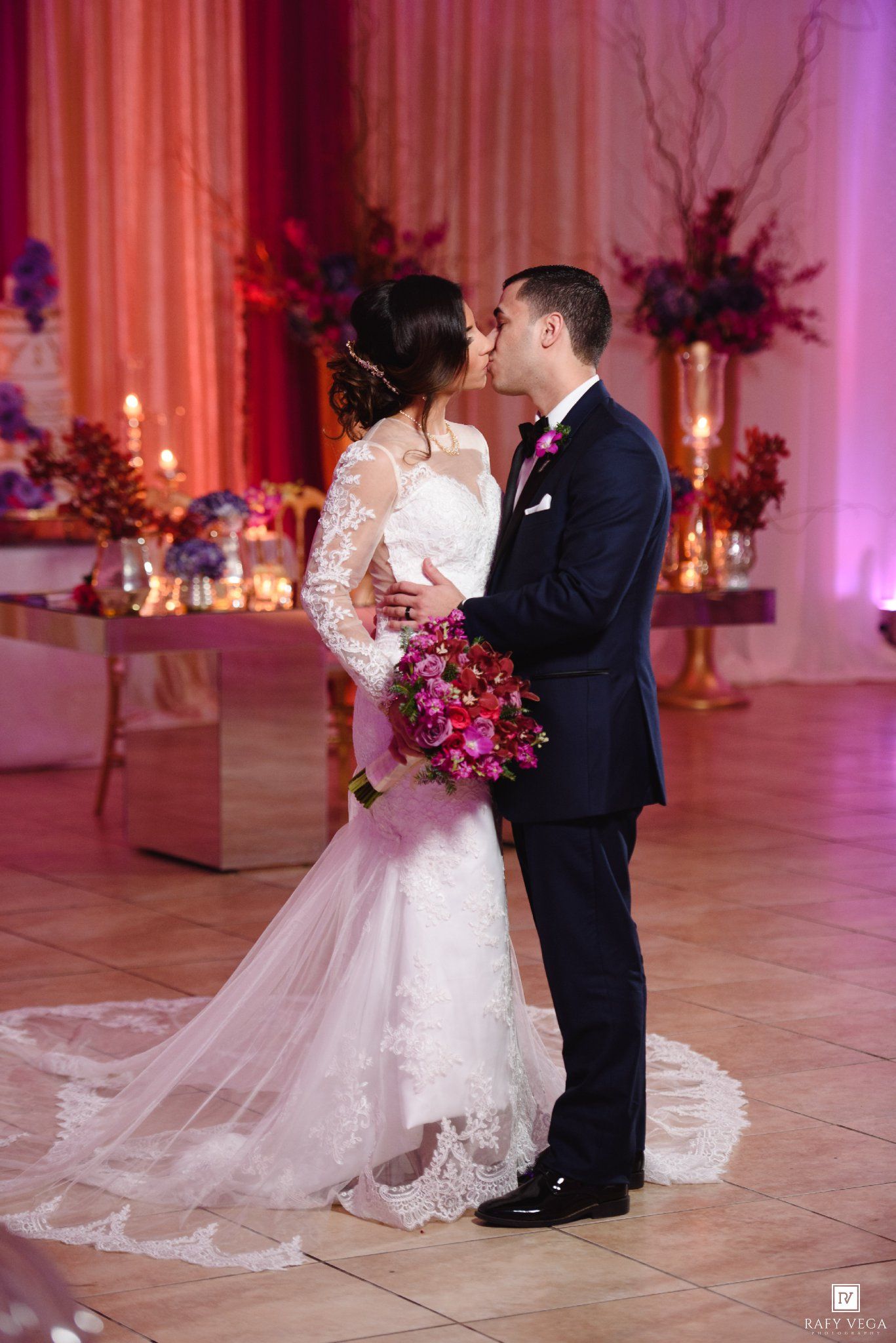 Bride and groom share a kiss on the dance floor, surrounded by floral arrangements and candlelight at their reception.