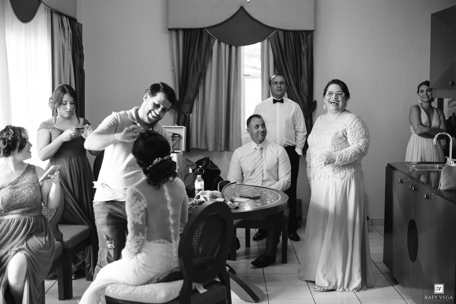Black and white wedding photo of bride and groom with guests gathered in elegant room during pre-ceremony preparations.
