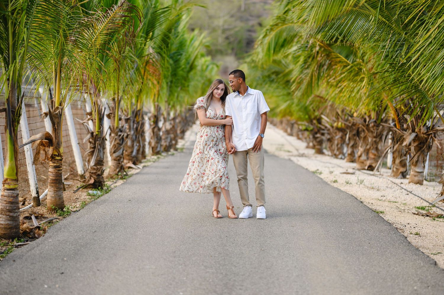 Couple in casual summer outfits embrace while walking along a paved path lined with tall palm trees at a tropical destination.