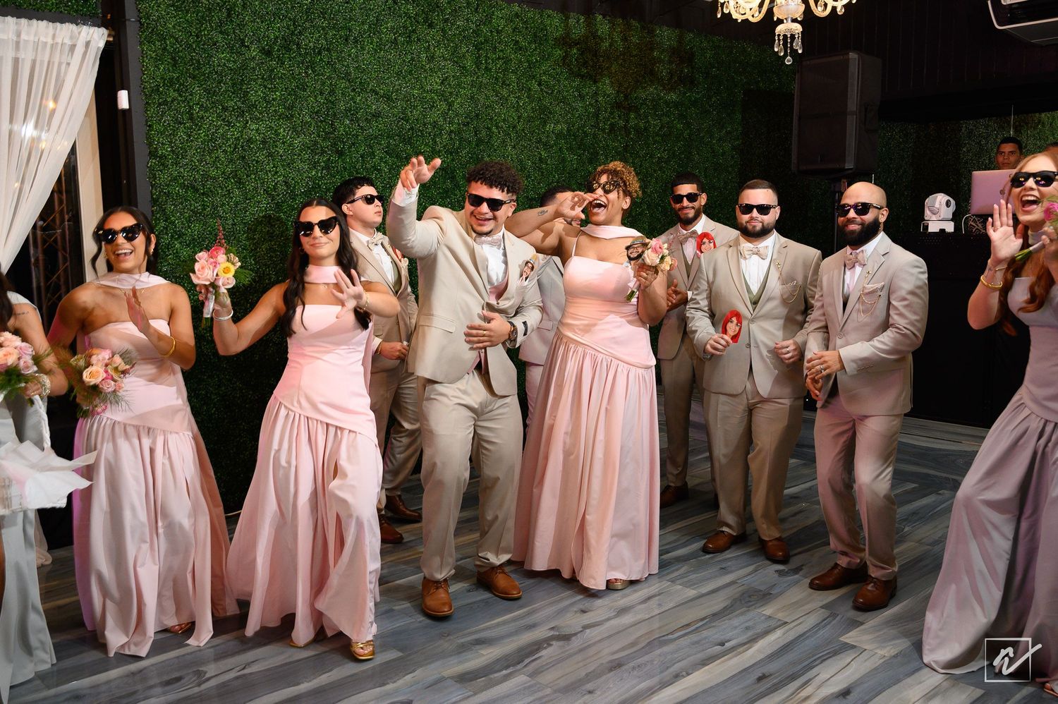 Bridesmaids in blush pink dresses and groomsmen in tan suits and sunglasses dance joyfully on a wedding reception dance floor.