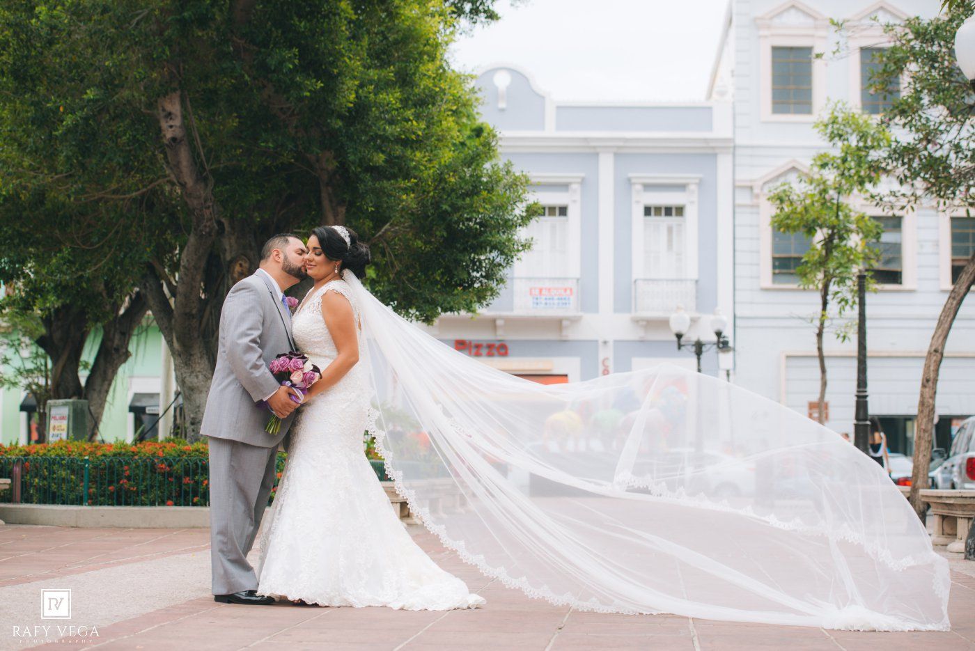 Bride and groom share a romantic kiss outdoors, with brides long veil flowing in the wind near a town square.