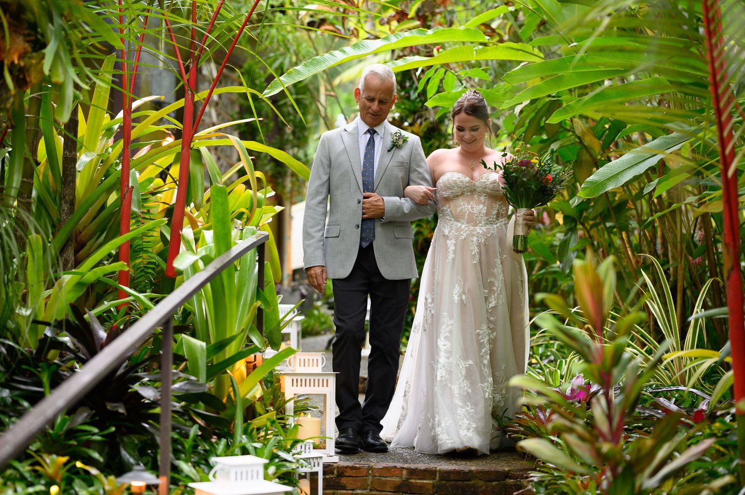 Bride in white gown walks with older man through lush tropical garden during outdoor wedding ceremony.