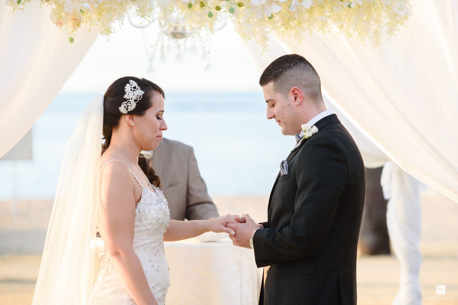 Bride and groom exchange rings during an outdoor beach wedding ceremony under a white floral arch.