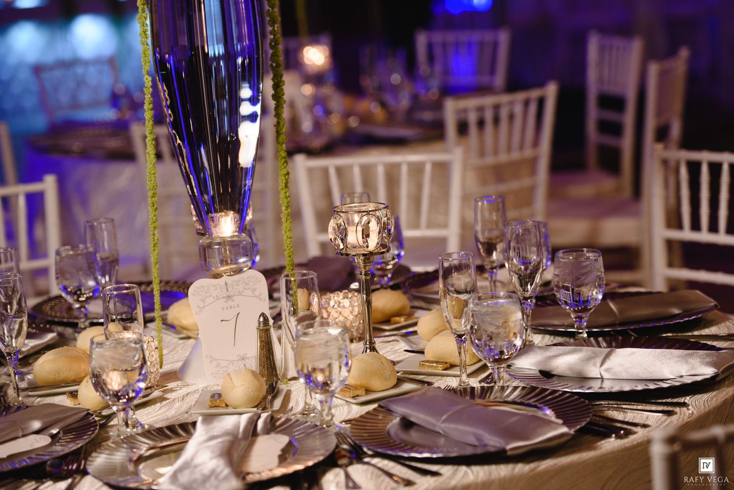 Elegant wedding reception table setting with silver charger plates, crystal glasses, and tall blue centerpieces.
