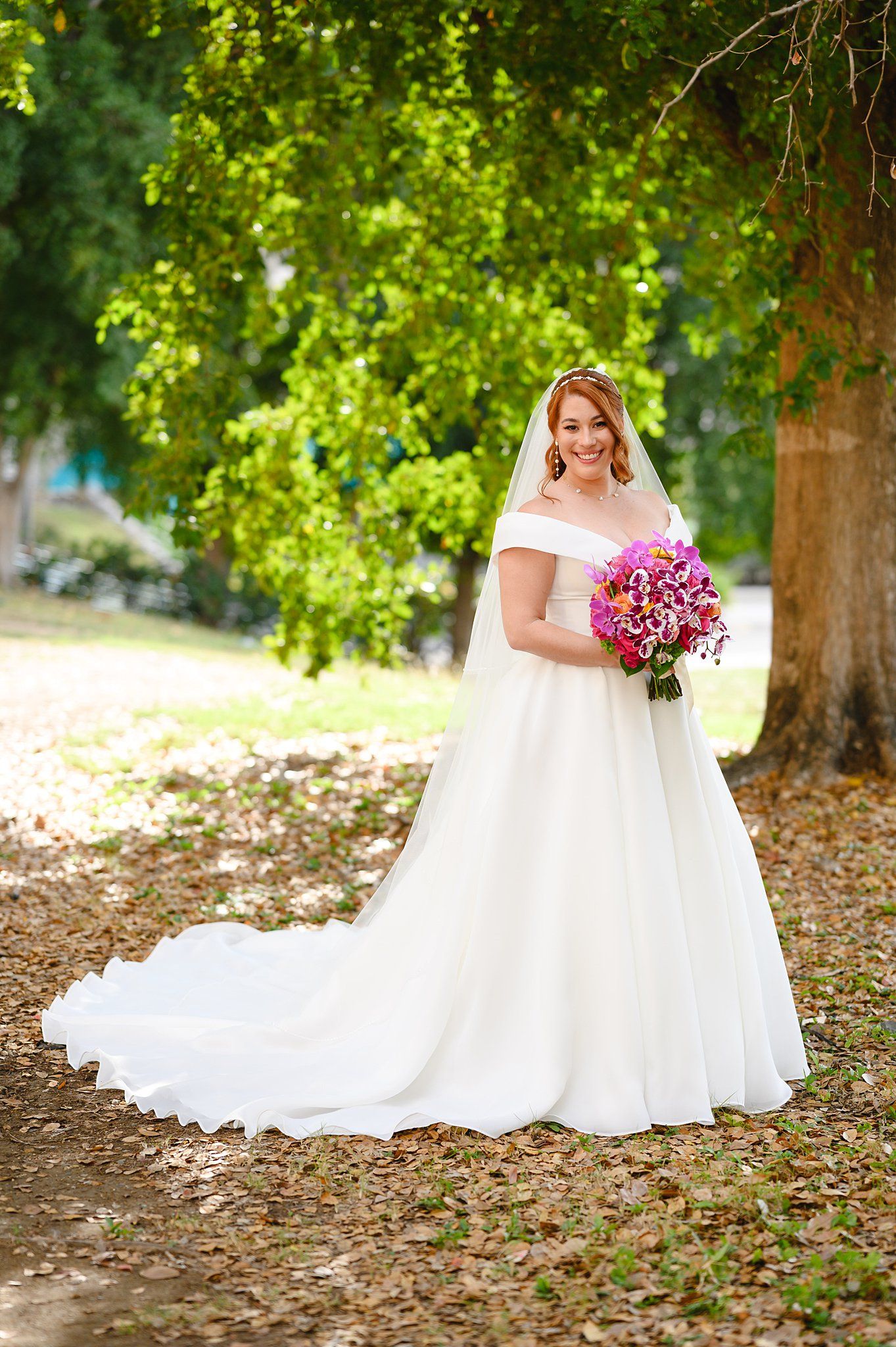 Bride in white off-shoulder gown with veil holds pink bouquet, standing under green trees on leaf-covered ground.