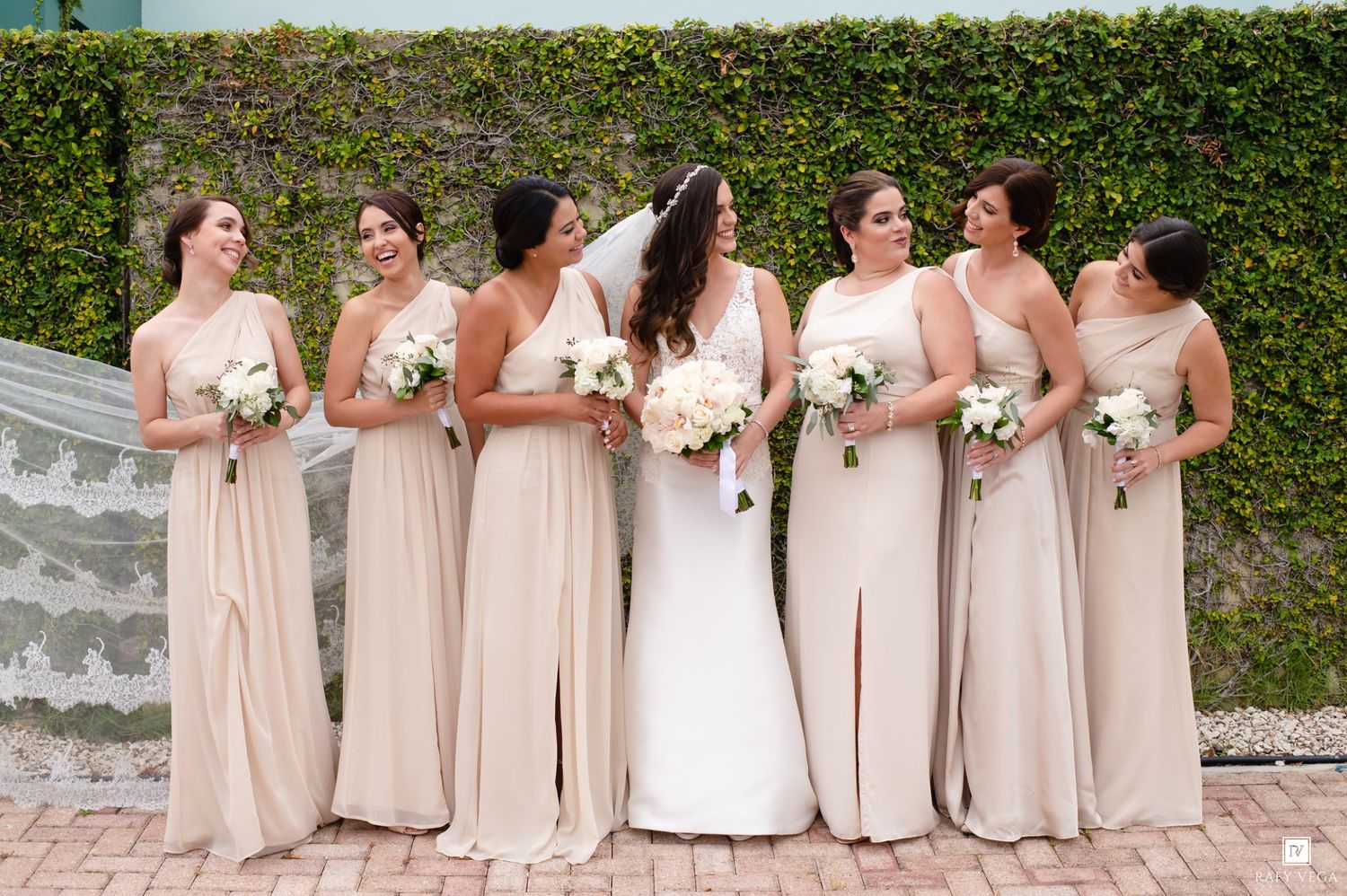 Bride and five bridesmaids in champagne gowns laughing together outdoors against a lush green ivy-covered wall.