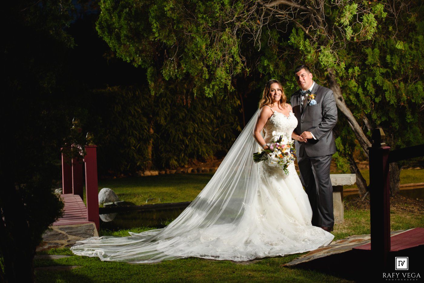 Bride and groom posing outdoors at night, bride wearing a long veil and white gown with a bouquet.