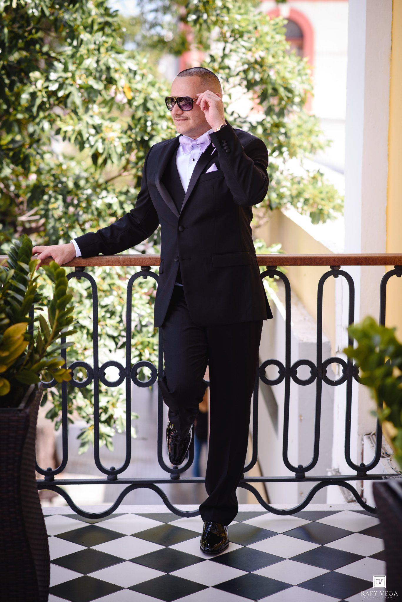 Elegant groom in black tuxedo and sunglasses posing on ornate balcony with lush greenery backdrop.