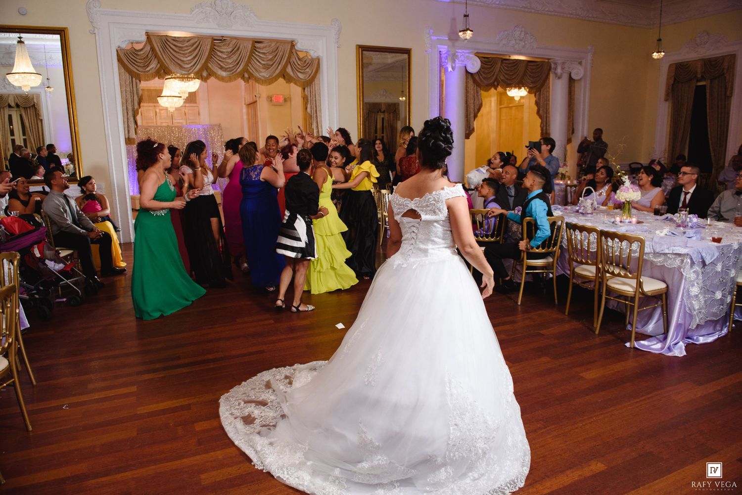 Bride in elegant white gown with train stands on dance floor surrounded by colorful guests at wedding reception.
