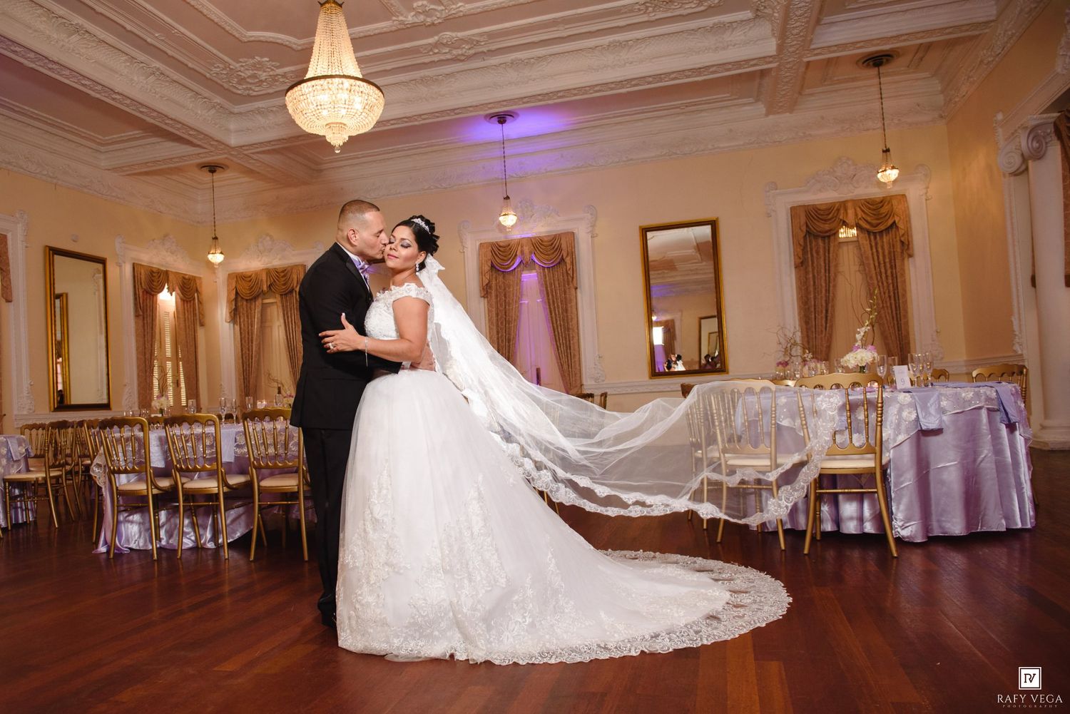 Bride and groom share romantic kiss in elegant ballroom with gold chairs and chandelier during wedding reception.