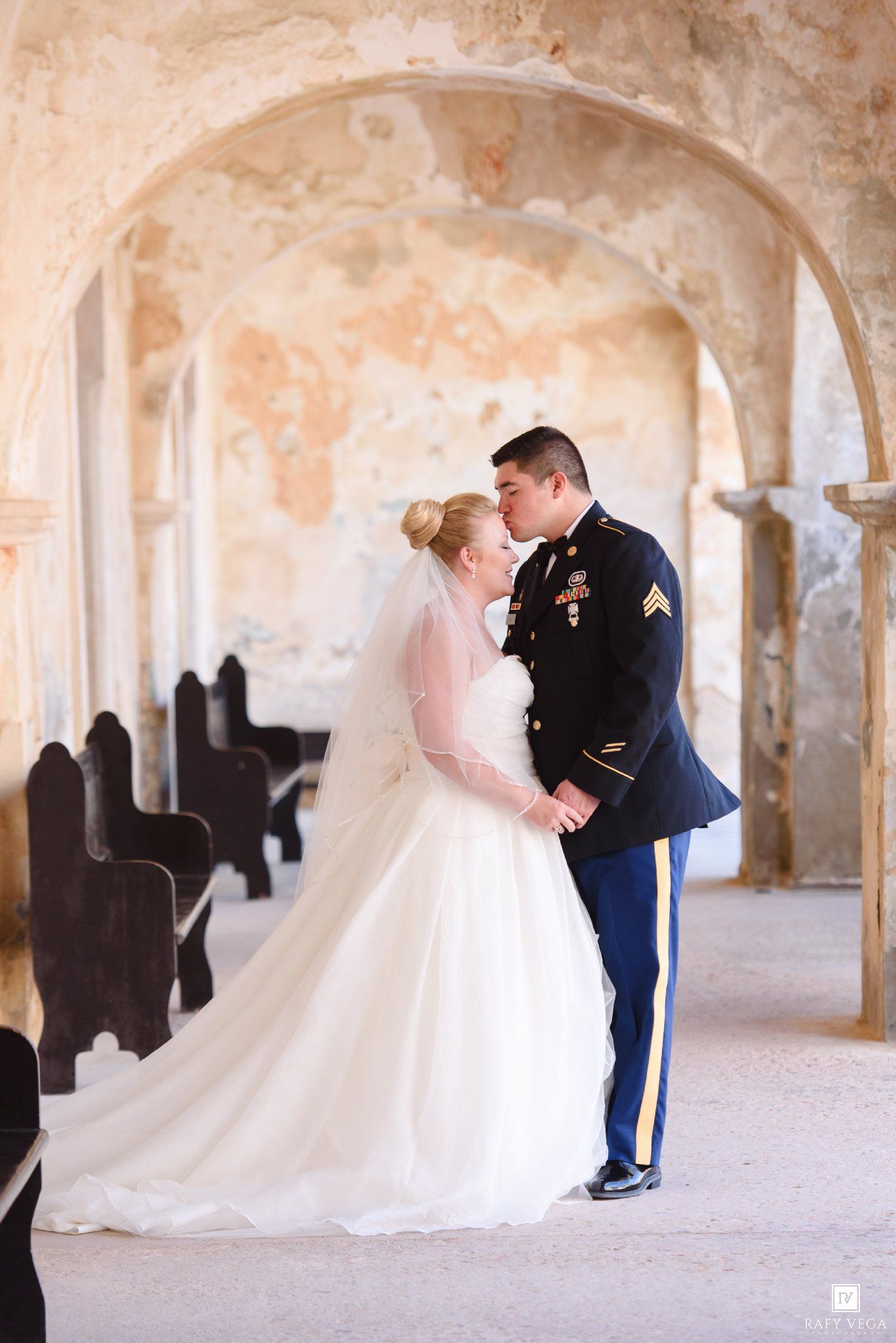 Military groom in dress uniform kissing bride in white gown under stone arch at historic wedding venue.
