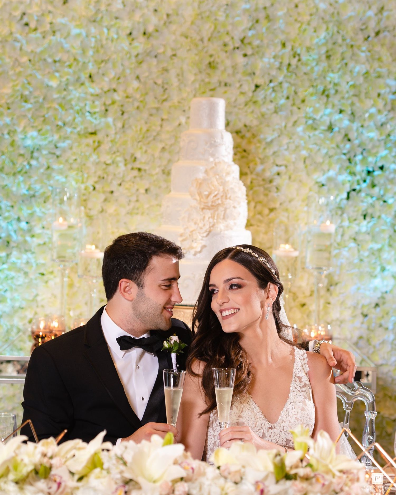 Bride and groom smiling at each other at sweetheart table with white floral wall and tiered wedding cake backdrop.