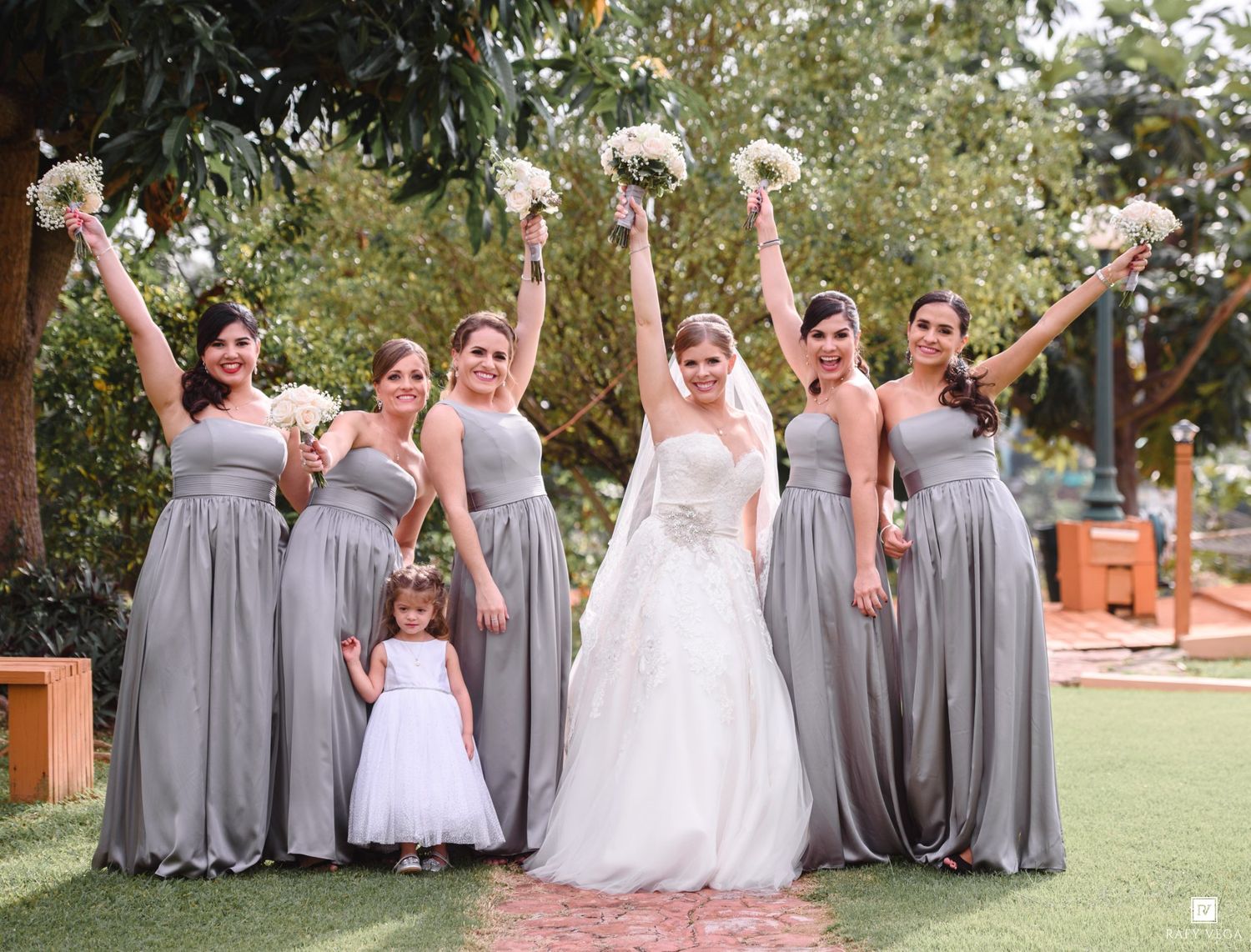 Bride and bridesmaids in gray dresses celebrate outdoors, raising bouquets with a flower girl in white.