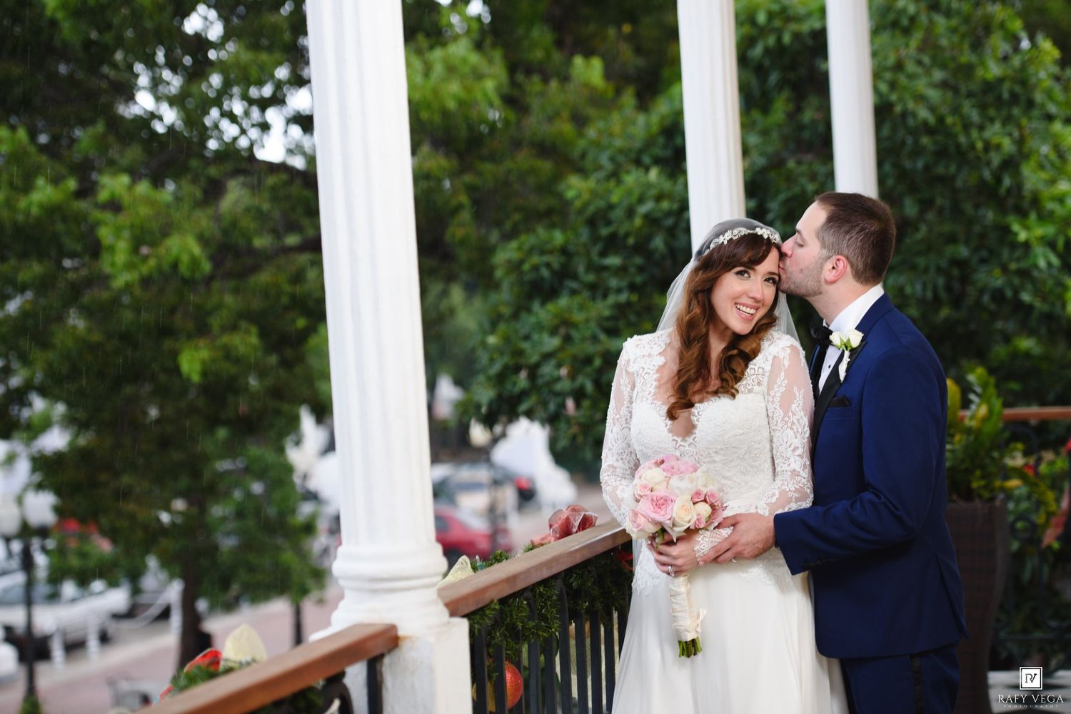 Bride and groom share a romantic moment on a white-columned balcony, bride holding a pink bouquet.