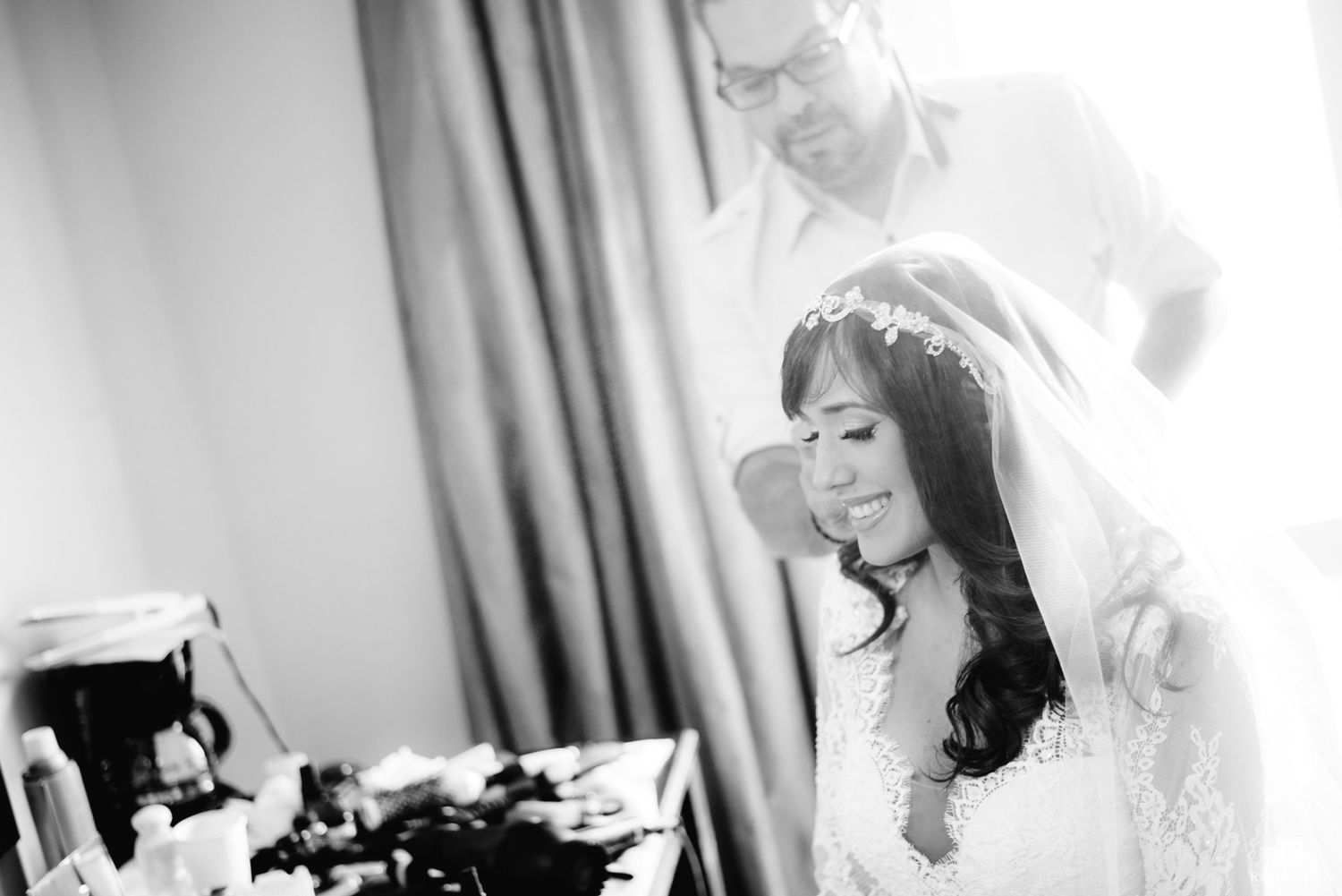 Bride in white gown smiles as groom adjusts her veil in black and white wedding photo.