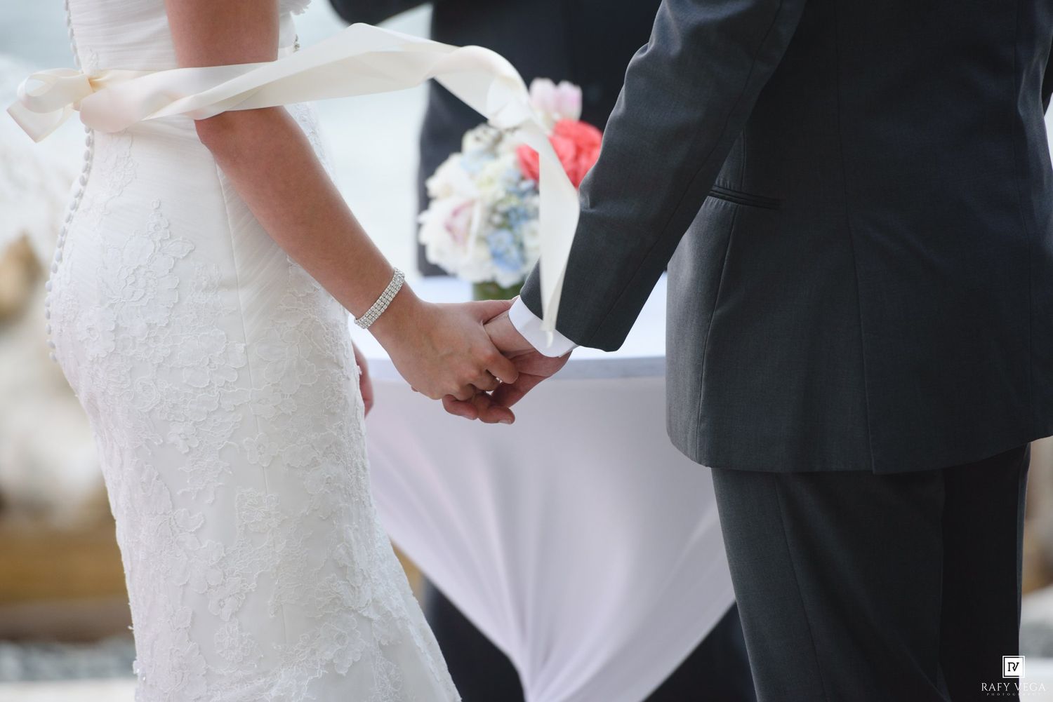 Bride and groom holding hands during wedding ceremony, bride in lace gown holding bouquet.