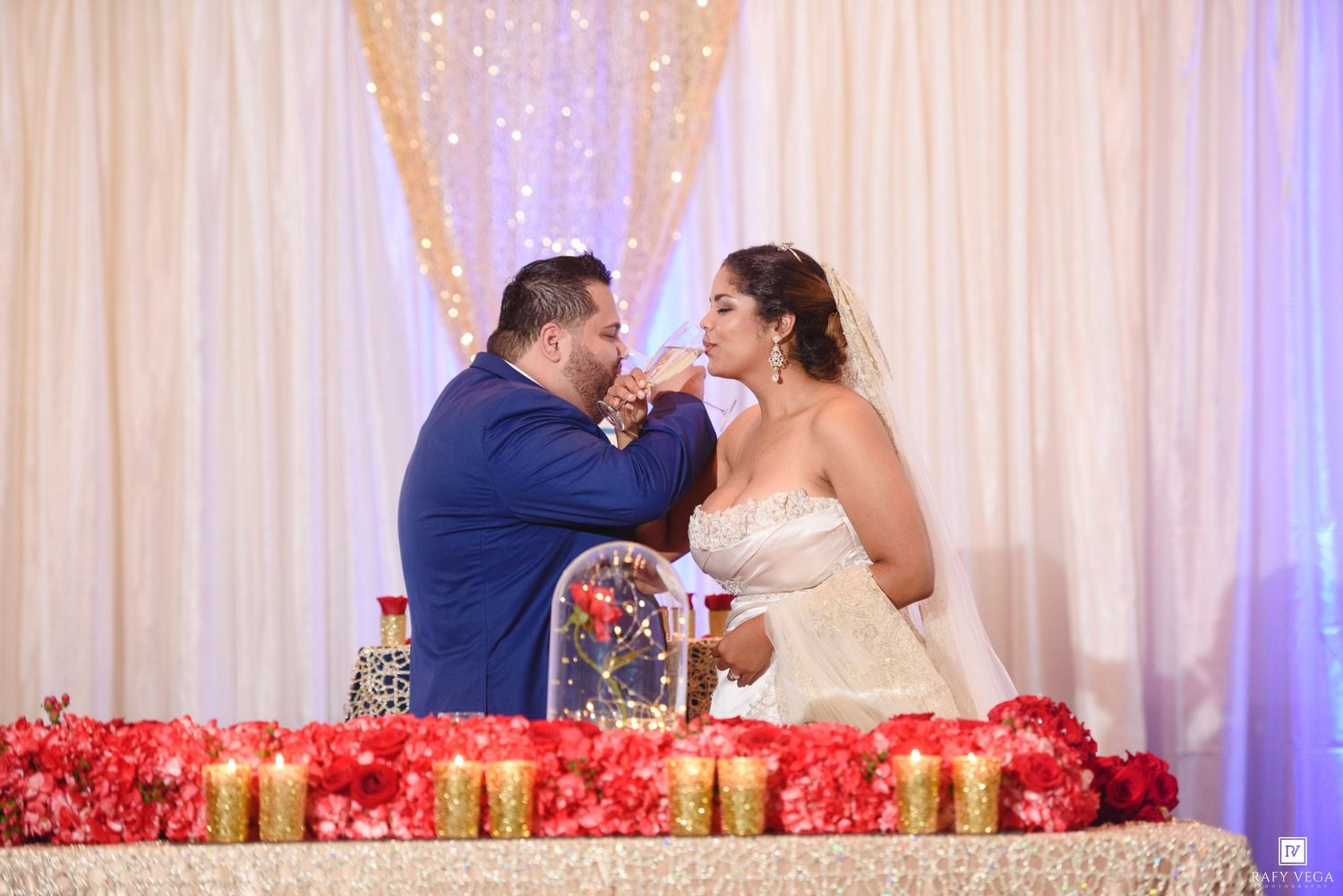 Groom in blue suit feeds wedding cake to bride in strapless gown at elegant reception with red floral arrangements.