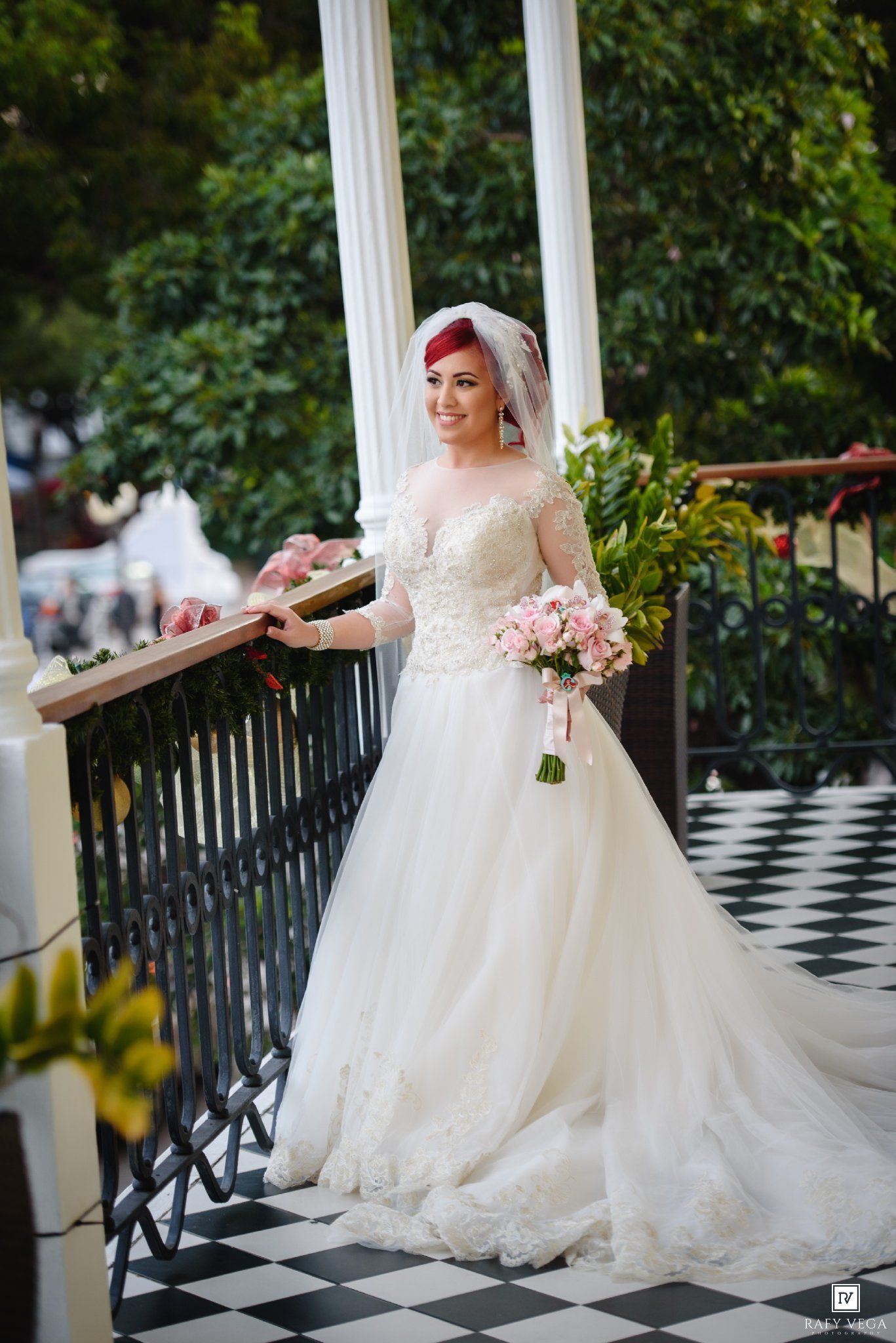 Bride in lace ball gown wedding dress holding pink bouquet on balcony with white columns and greenery.
