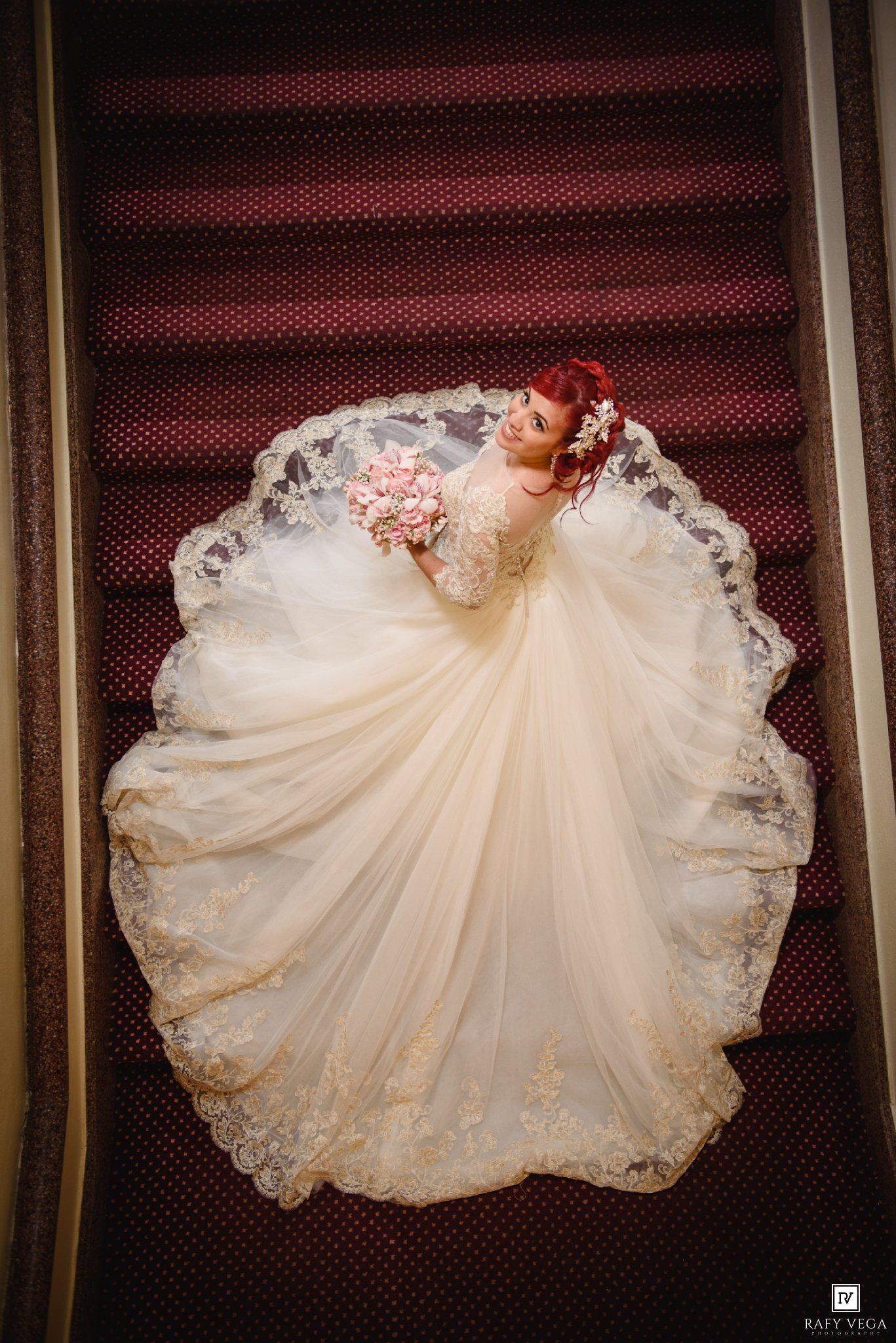 Bride in ivory ball gown with lace trim holds pink bouquet on stairs, photographed from above.