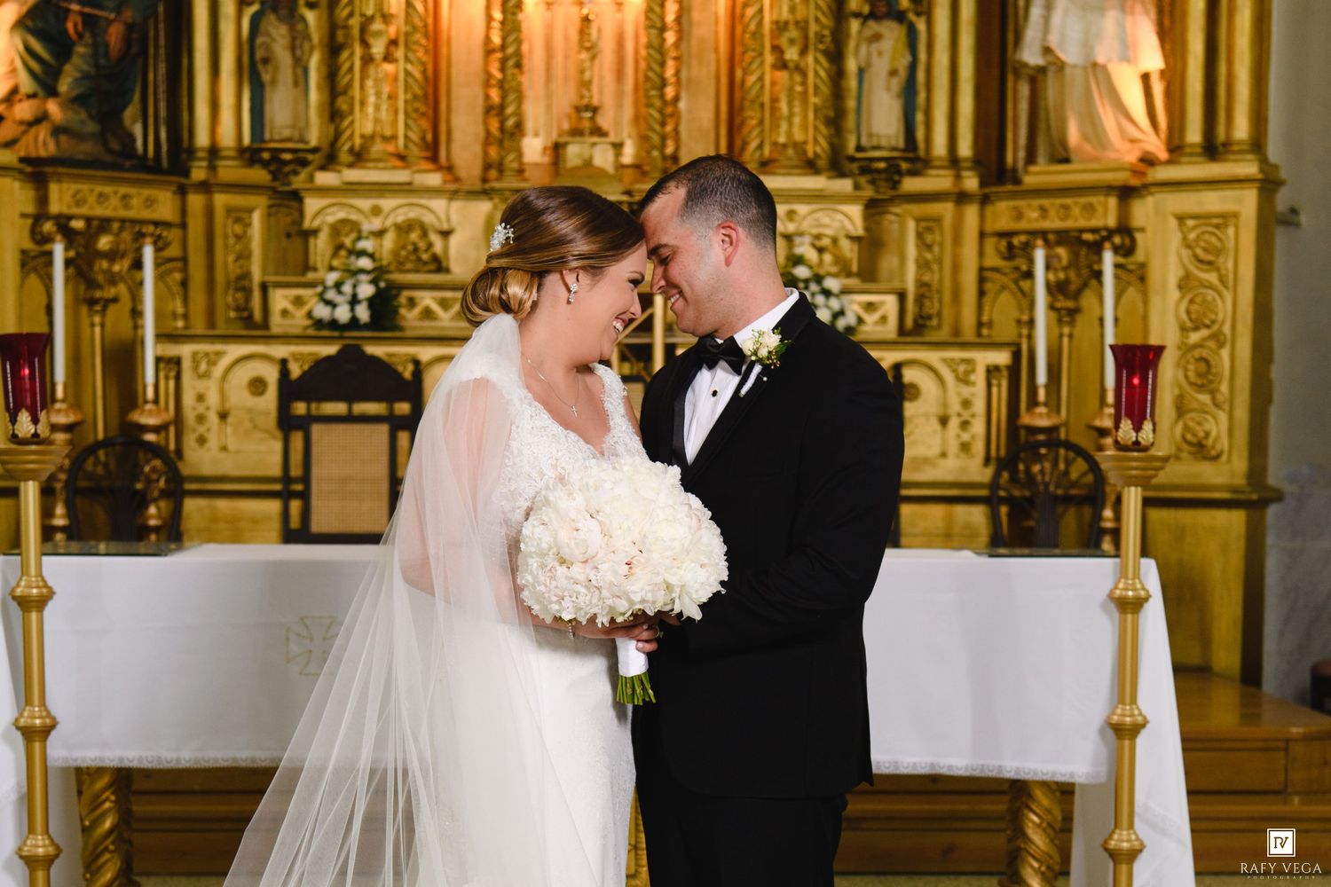 Bride and groom share an intimate moment at a church altar, bride holding white bouquet in elegant wedding attire.