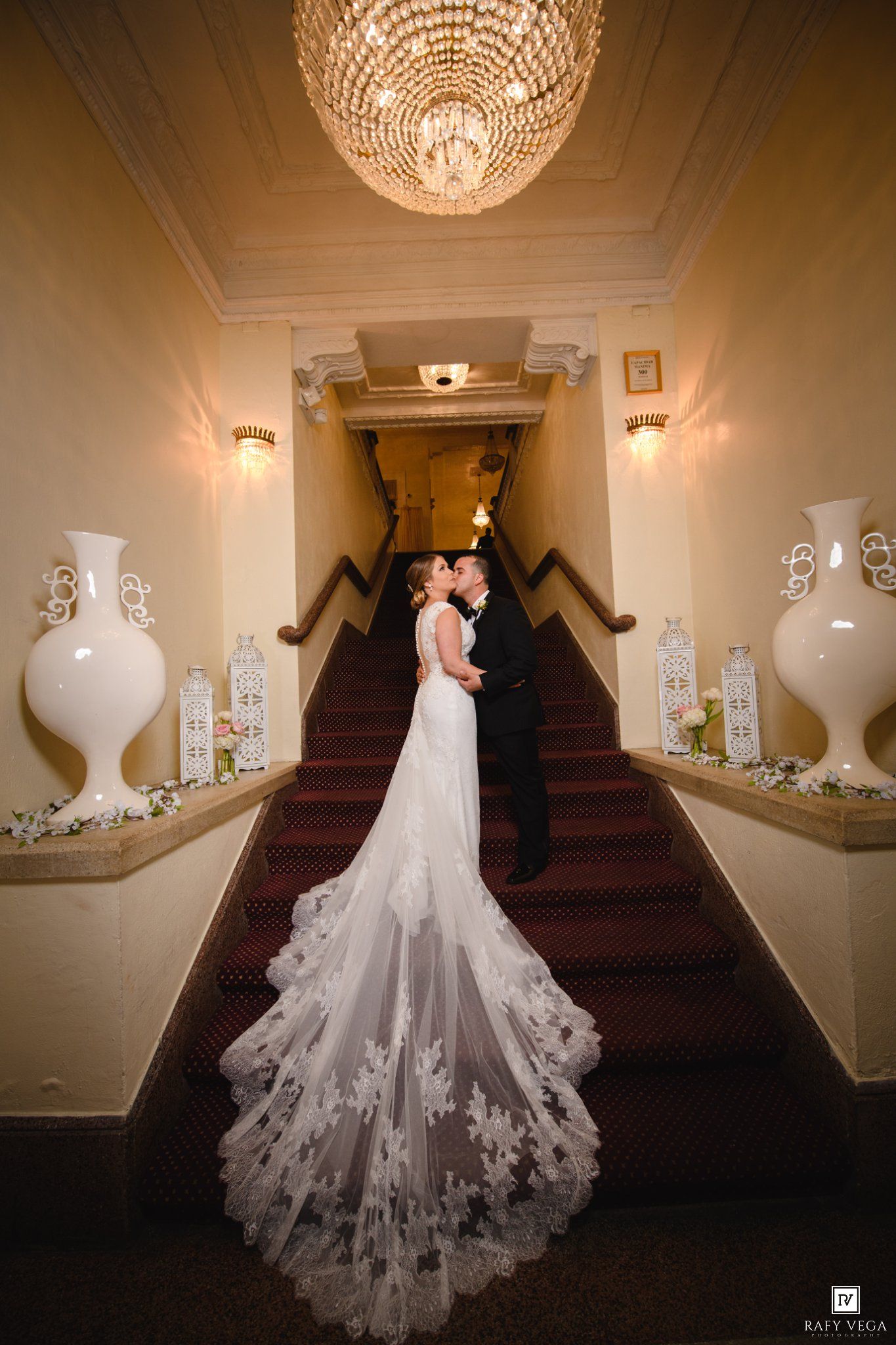 Bride and groom share a romantic kiss on elegant staircase with crystal chandelier and white vases at wedding venue.