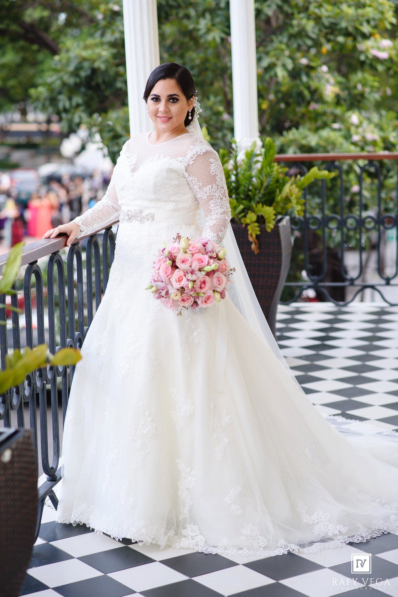 Bride in white lace ball gown with long sleeves holds pink bouquet on checkered floor balcony.