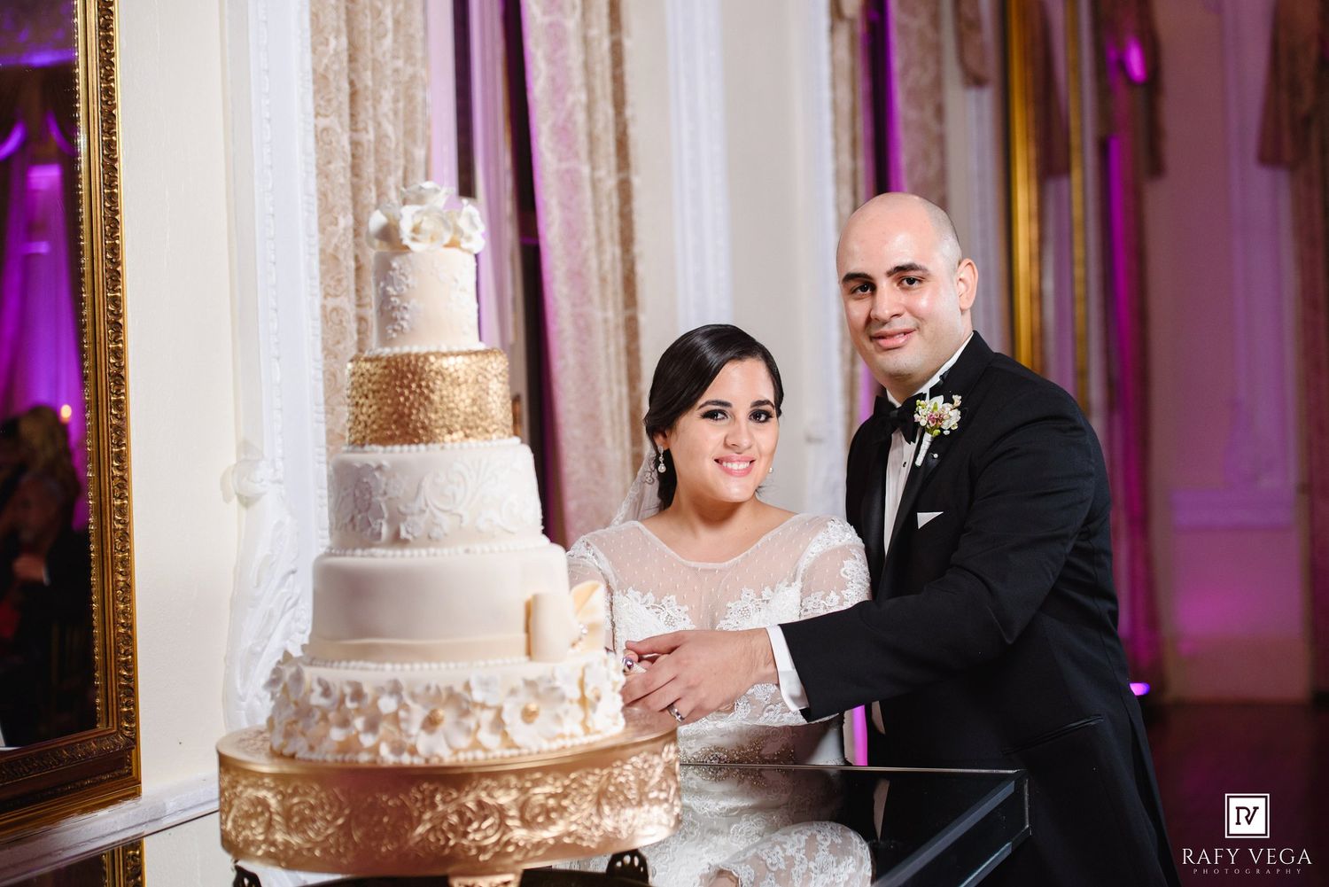 Bride and groom cutting elegant five-tier white and gold wedding cake at luxury ballroom reception.