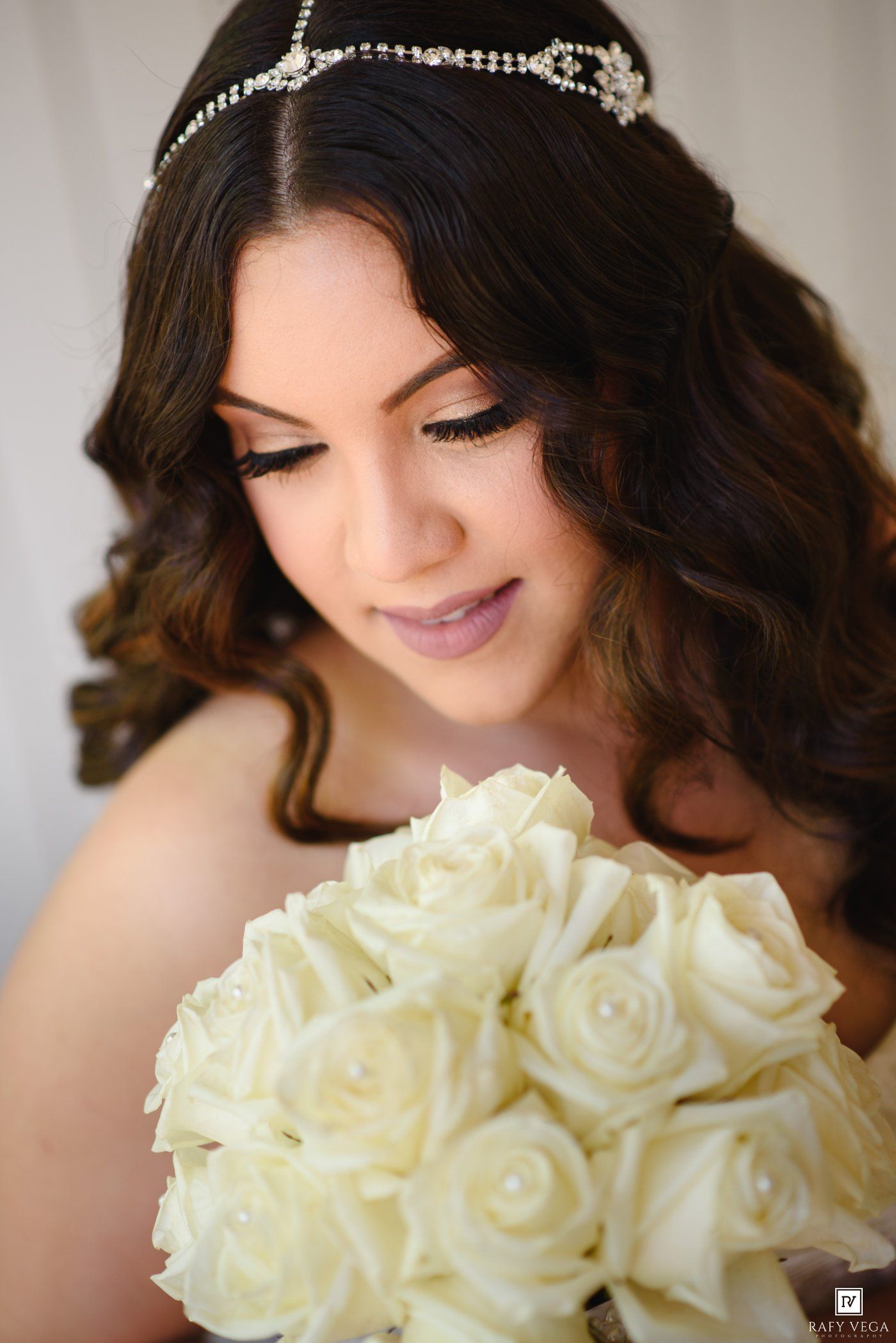 Bride with dark curly hair wearing a crystal tiara holds a bouquet of white roses, looking down gracefully.