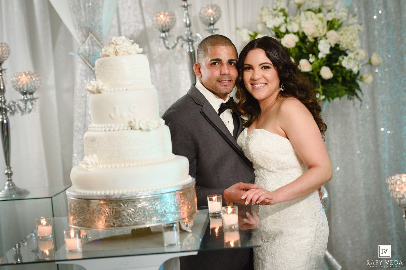 Bride and groom posing beside elegant white four-tier wedding cake with candles at their wedding reception.