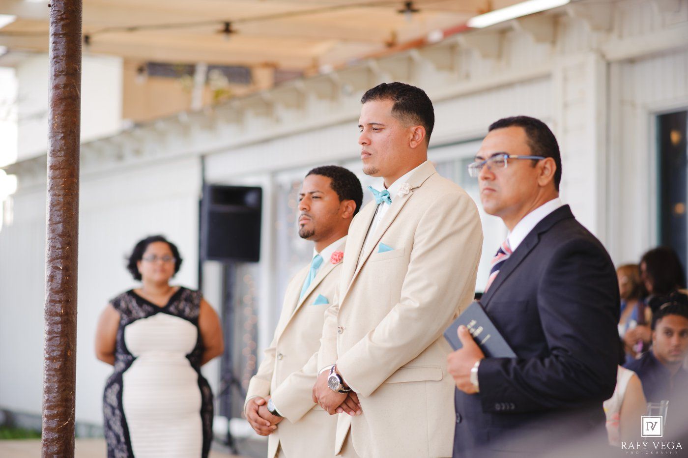 Three men in suits stand attentively during an outdoor wedding ceremony, with guests seated in the background.