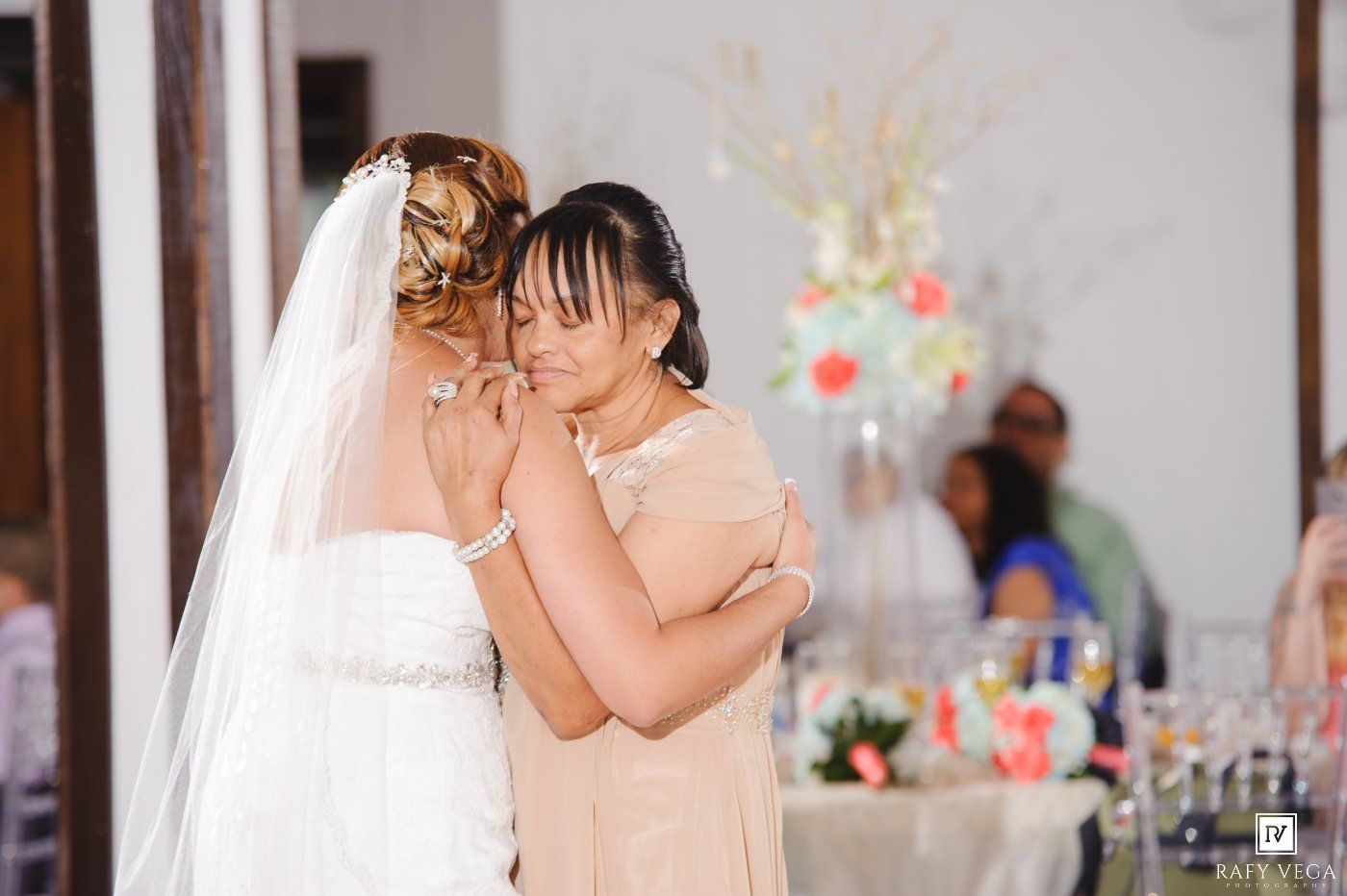 Bride in white gown and veil shares emotional moment with guest in champagne dress at elegant wedding reception.