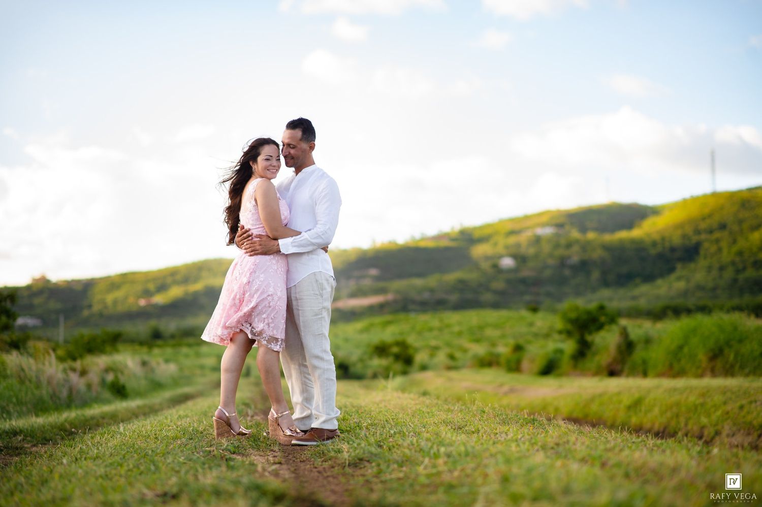 Couple embracing on a scenic green hillside path, woman in pink dress, man in white outfit, under a blue sky.