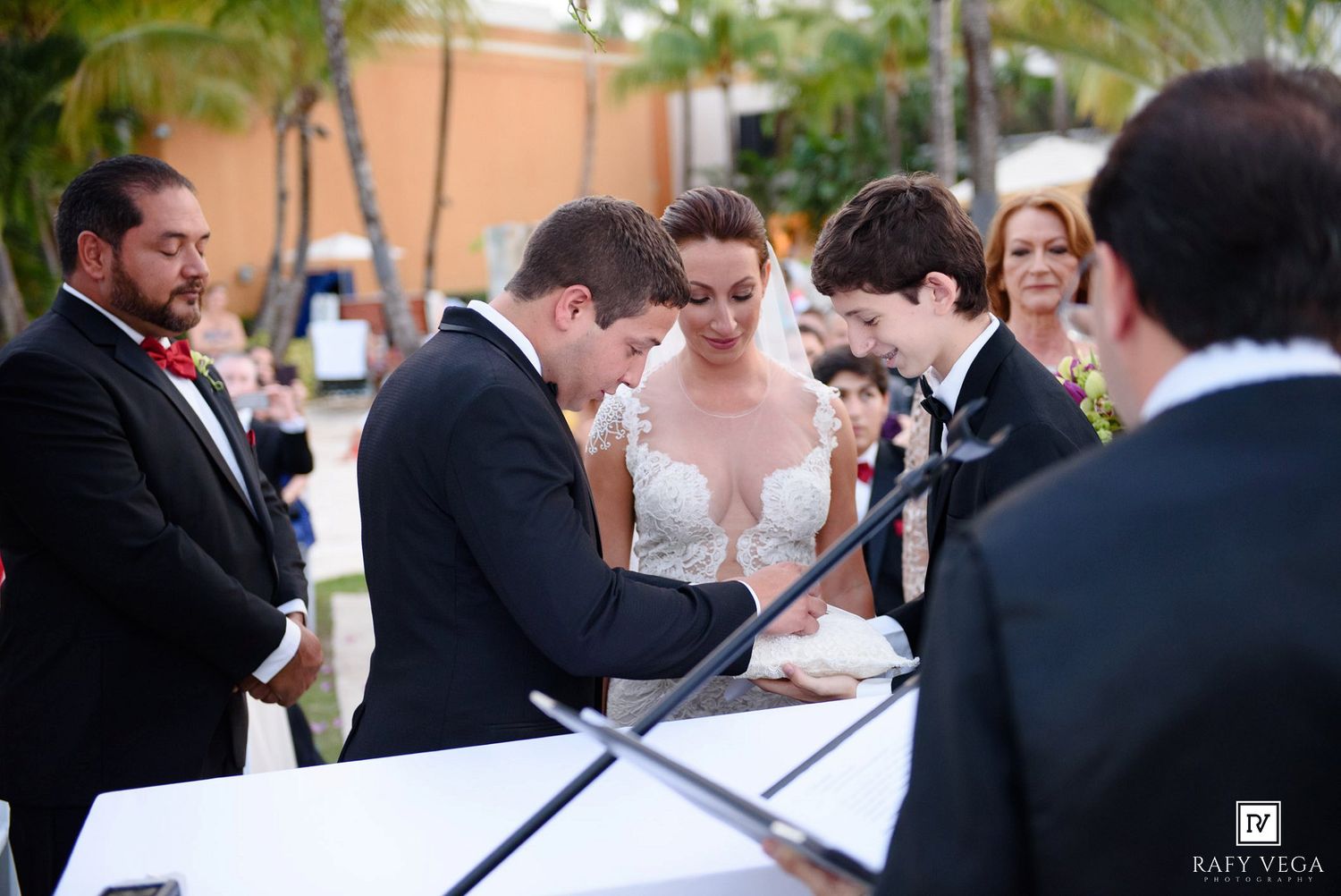 Bride and groom signing wedding documents during outdoor ceremony with guests and officiant watching.
