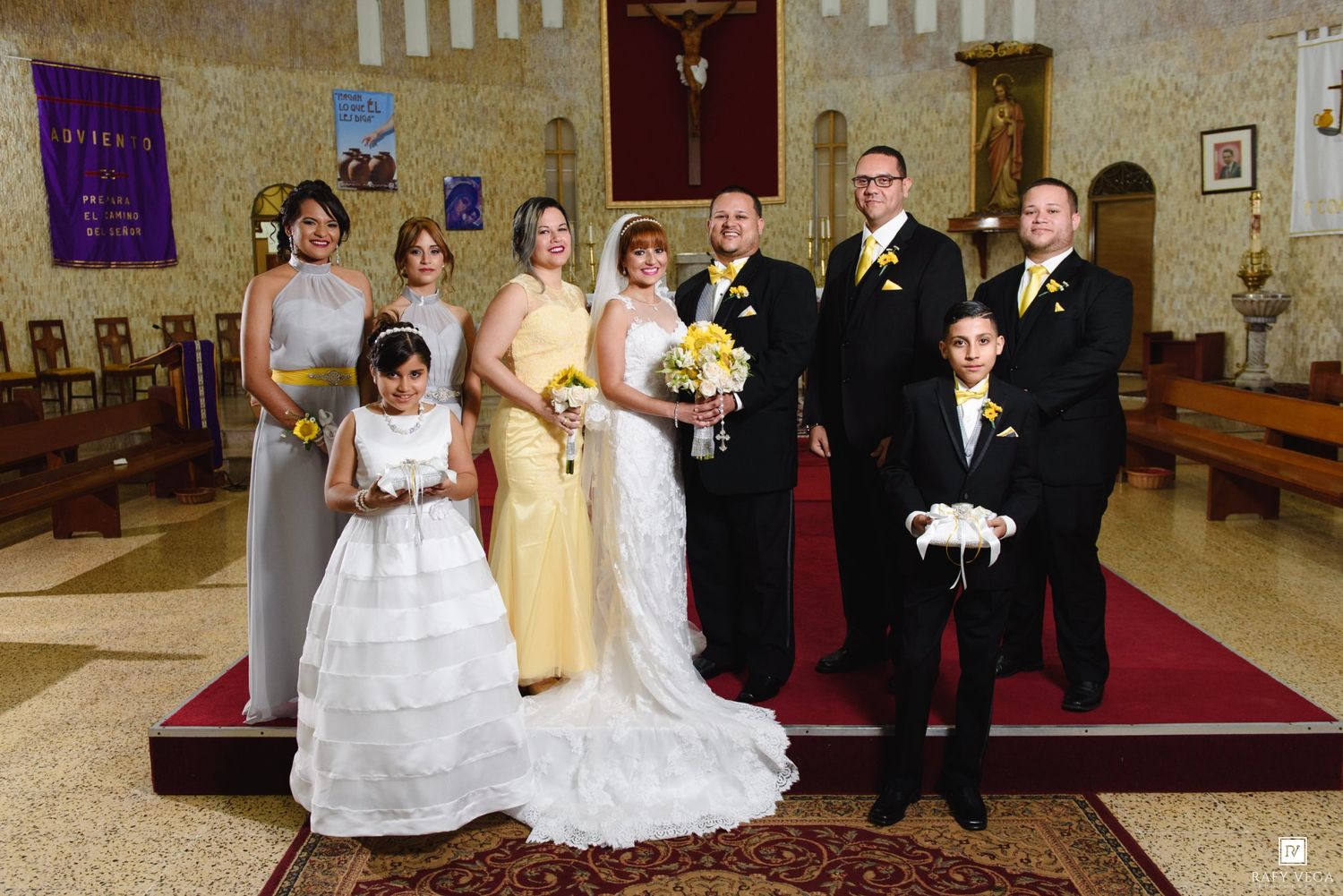 Wedding party group portrait in church with bride, groom, bridesmaids in yellow, groomsmen, and flower girl.
