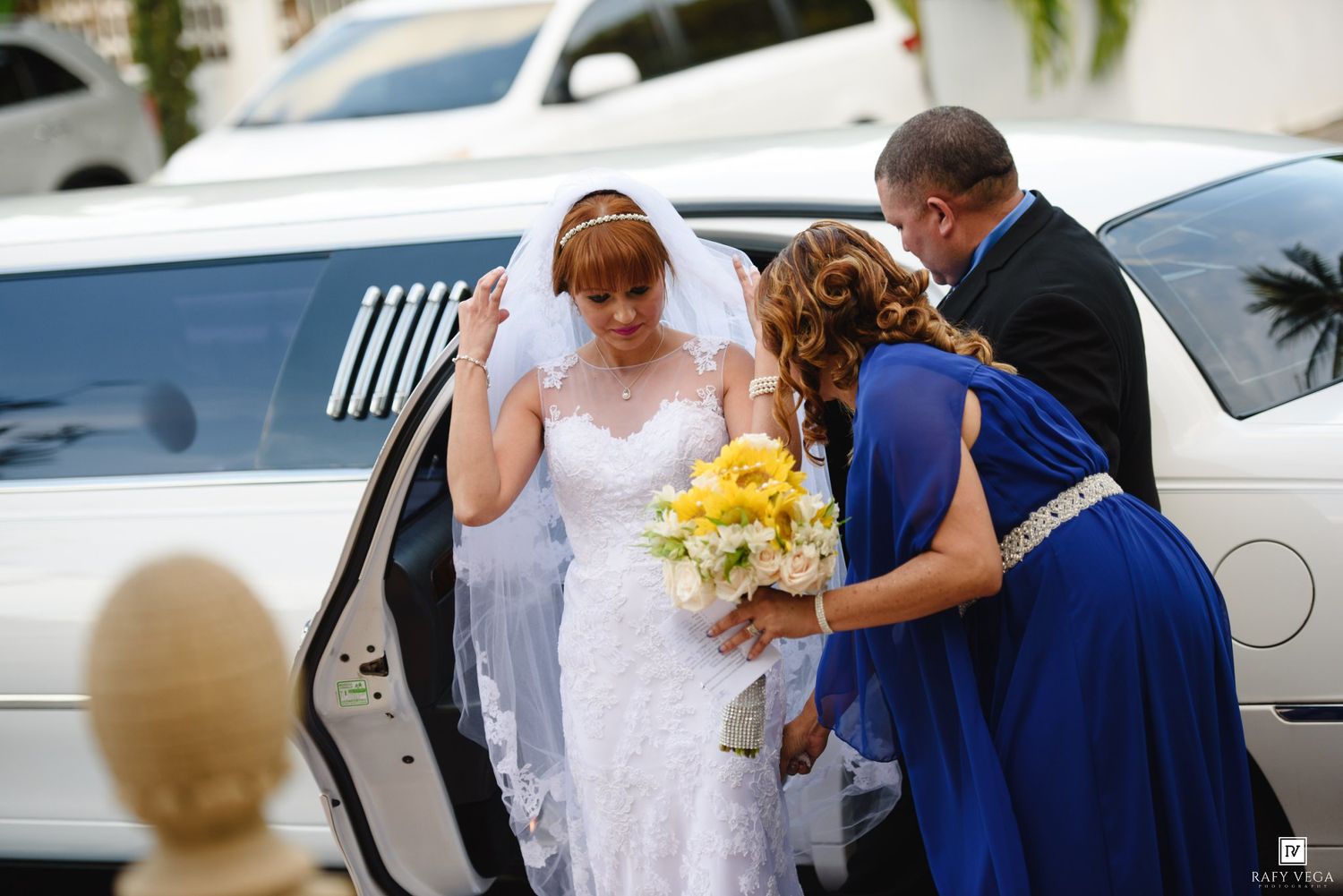 Bride in white lace gown holding yellow bouquet being helped out of a white limousine by wedding attendants.