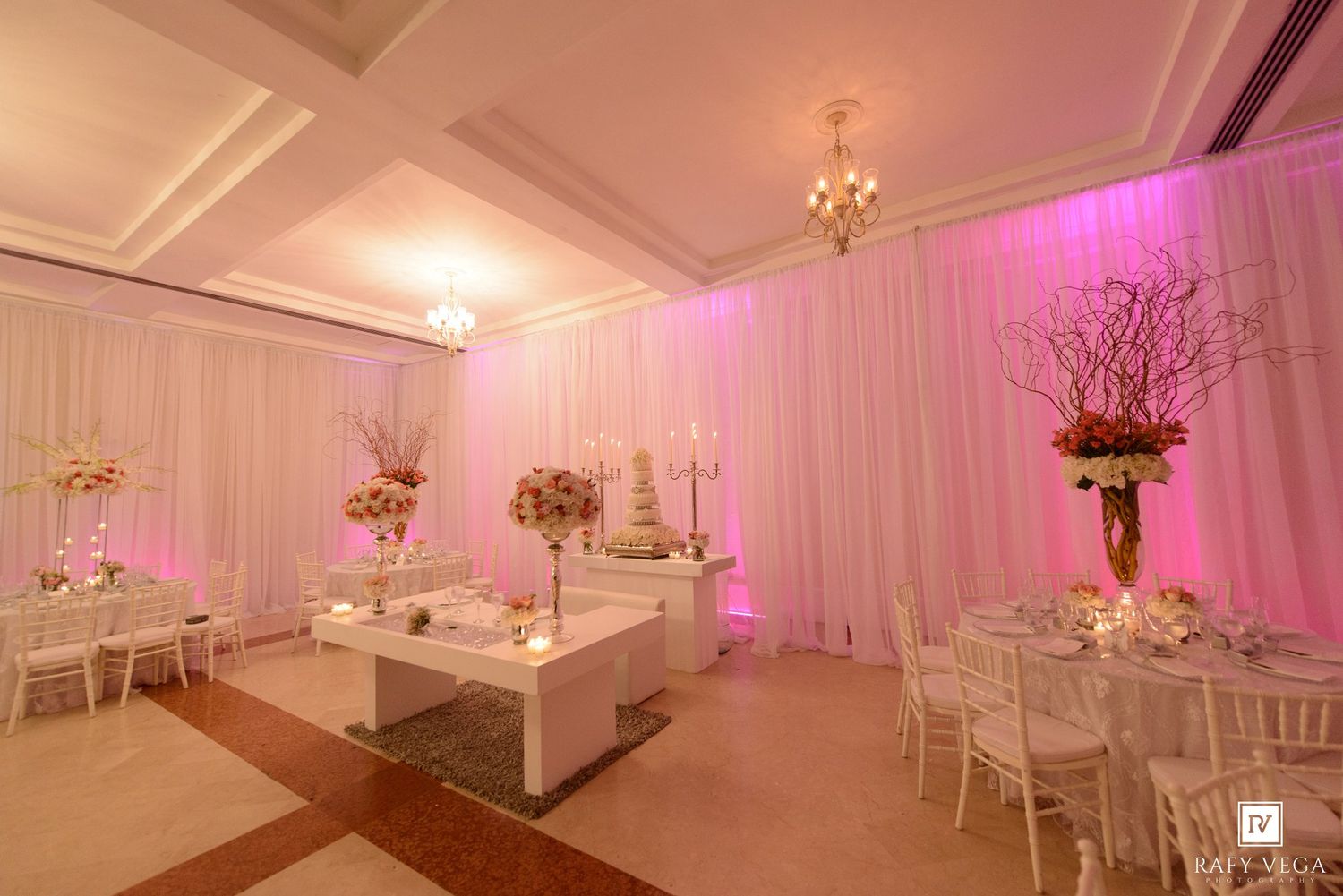 Elegant wedding reception hall with pink uplighting, white draping, floral centerpieces, and round tables with chiavari chairs.