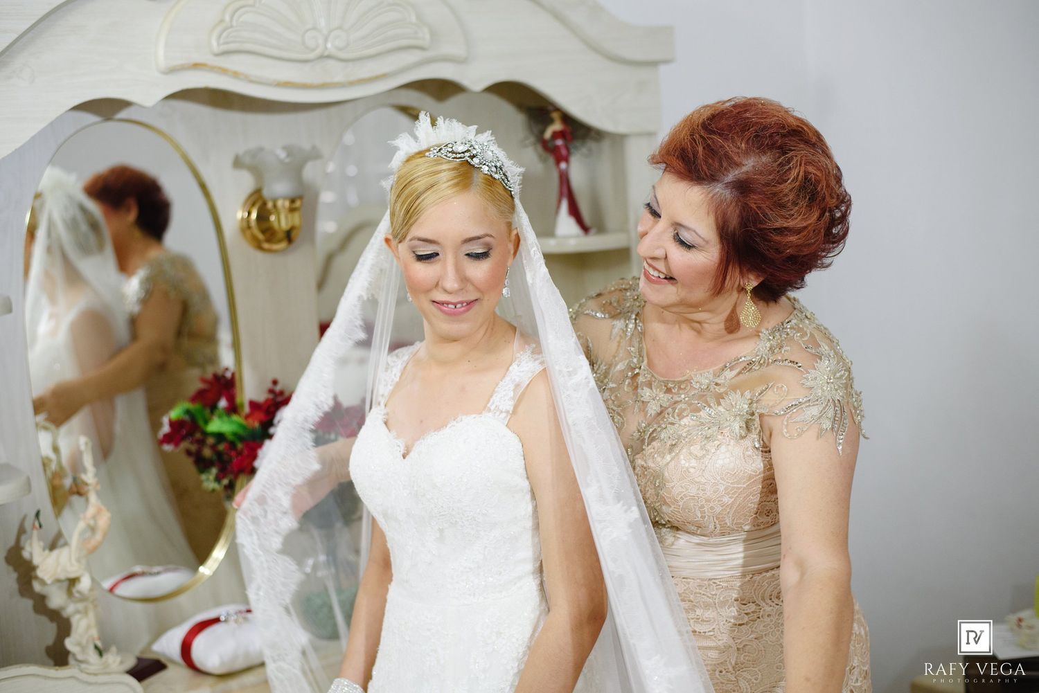 A bride in a white wedding gown and veil being helped by an older woman in a gold dress near a mirror.