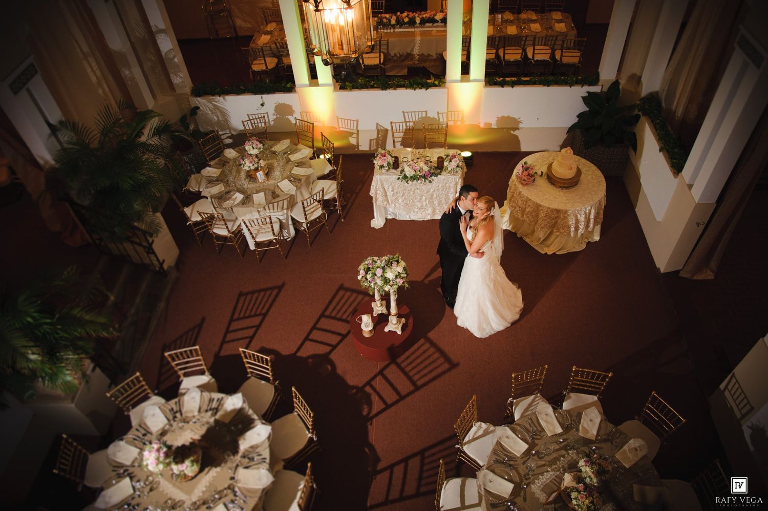 Aerial view of a wedding couple sharing a first dance surrounded by elegantly set round tables in a warmly lit reception hall.
