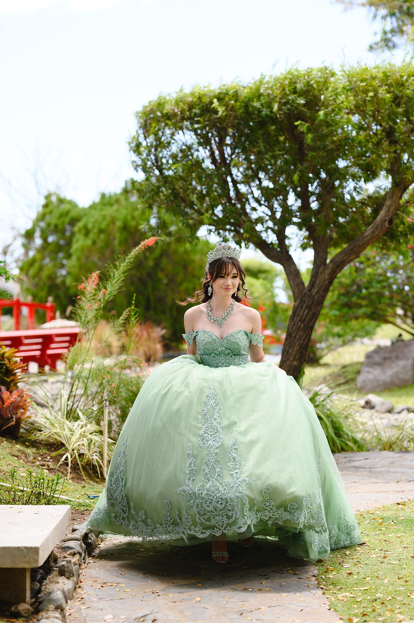 Young woman in elegant mint green quinceañera ballgown with lace details and tiara walking through a garden.