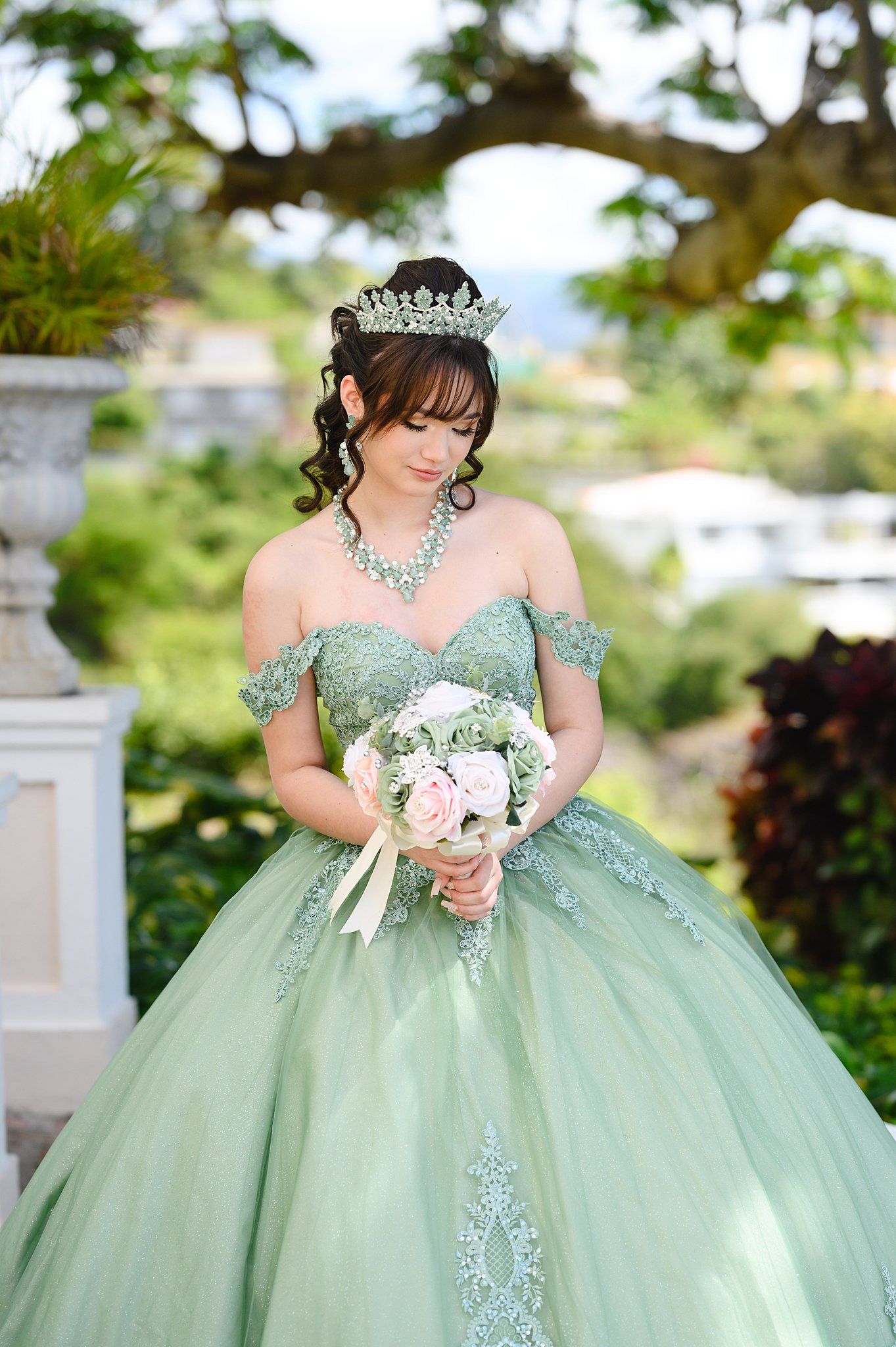 Young woman in sage green quinceañera ball gown with tiara holds pink bouquet in lush garden setting.