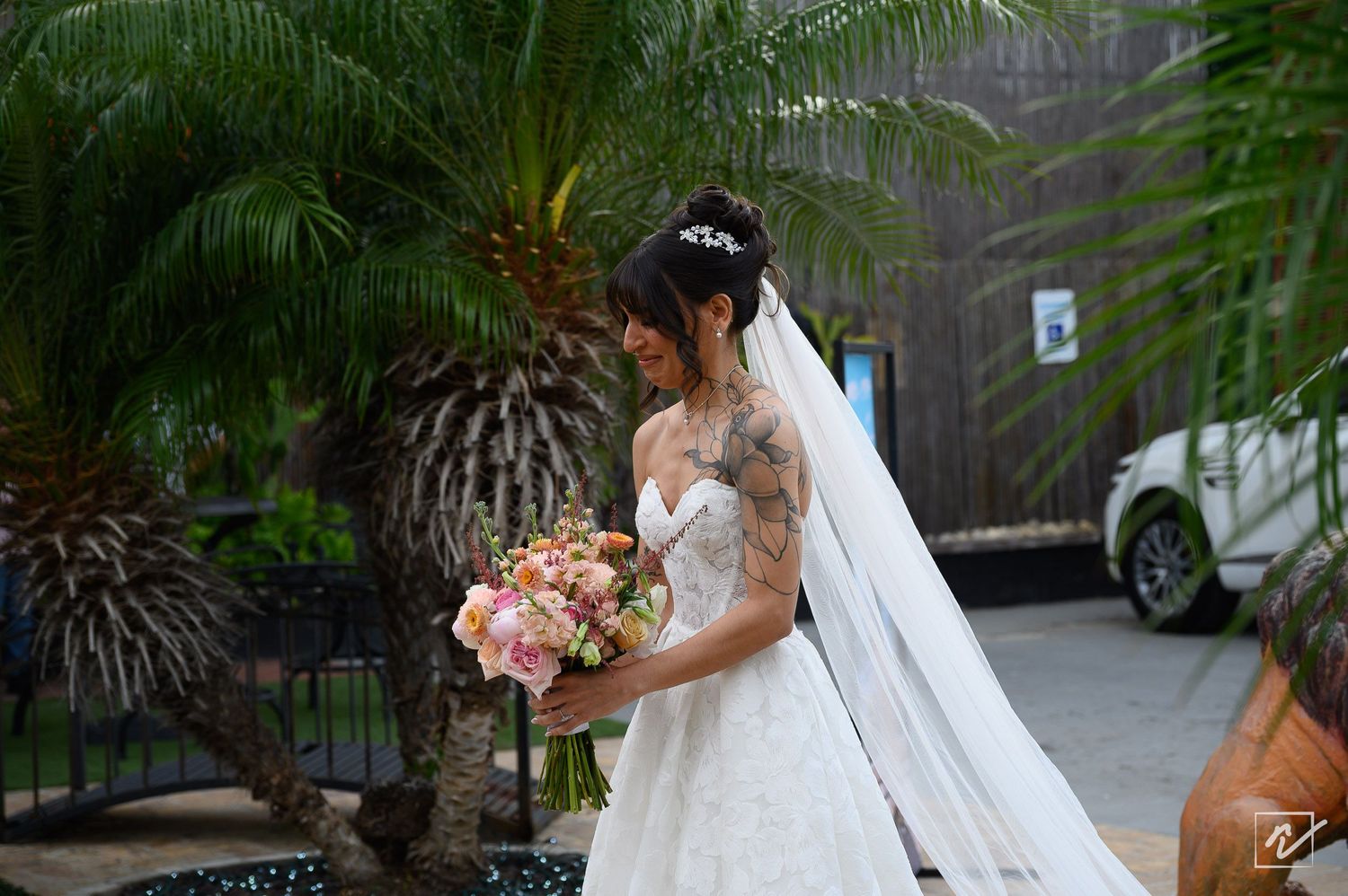 Bride in a white lace wedding gown with a long veil and tiara holds a pink floral bouquet beside palm trees outdoors.