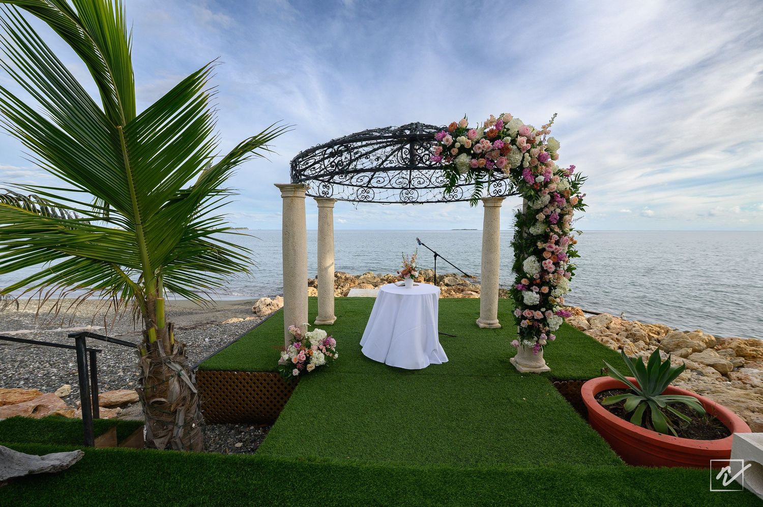 Oceanfront wedding gazebo with pink floral arch, white draped table, and artificial turf aisle beside a palm tree overlooking the sea.