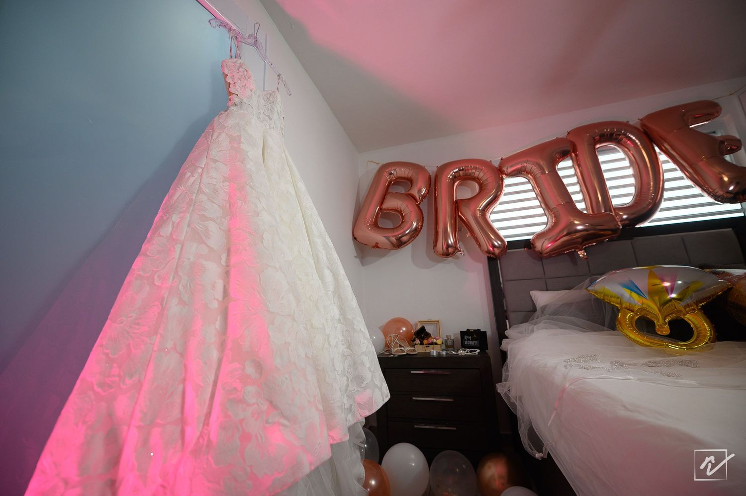 White lace wedding dress hanging in a bedroom with rose gold BRIDE letter balloons, a bed with pillows, and bridal party decorations.